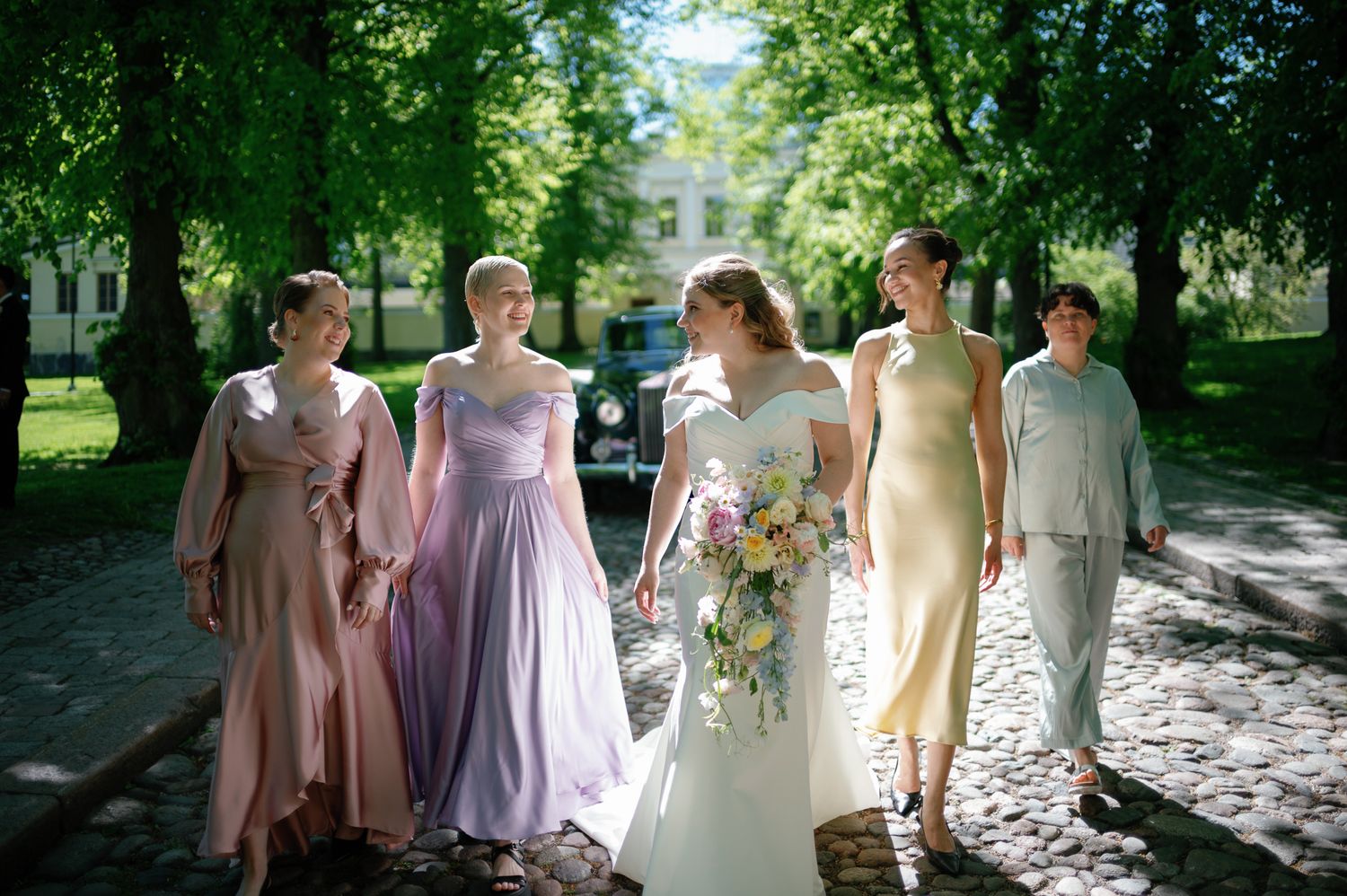 Bride in white gown walks with bridesmaids in pastel dresses, holding bouquet, on cobblestone path lined with green trees.