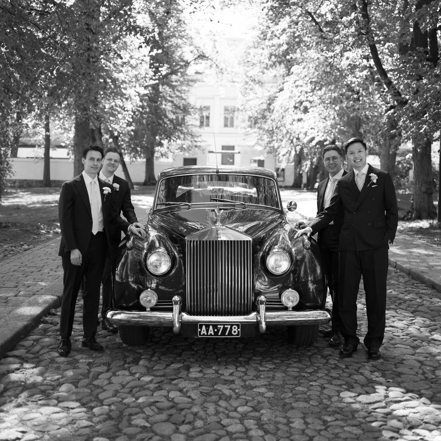 Black and white photo of three men in suits posing with a classic vintage Rolls-Royce on a cobblestone street.