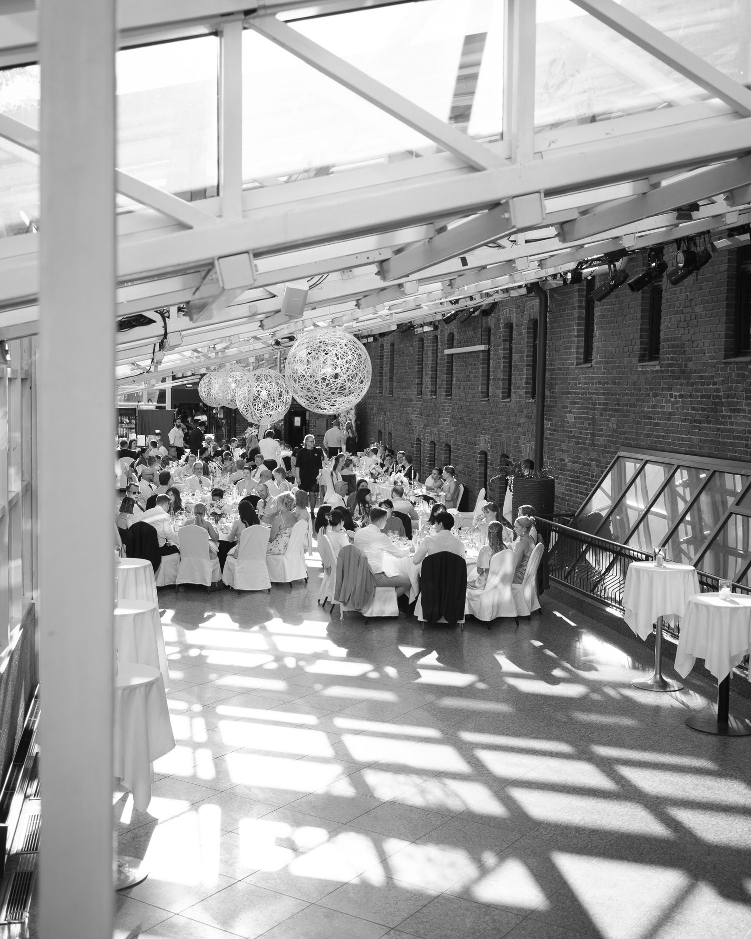 Black and white photo of an elegant banquet hall with guests seated at round tables under a glass ceiling with decorative chandeliers.