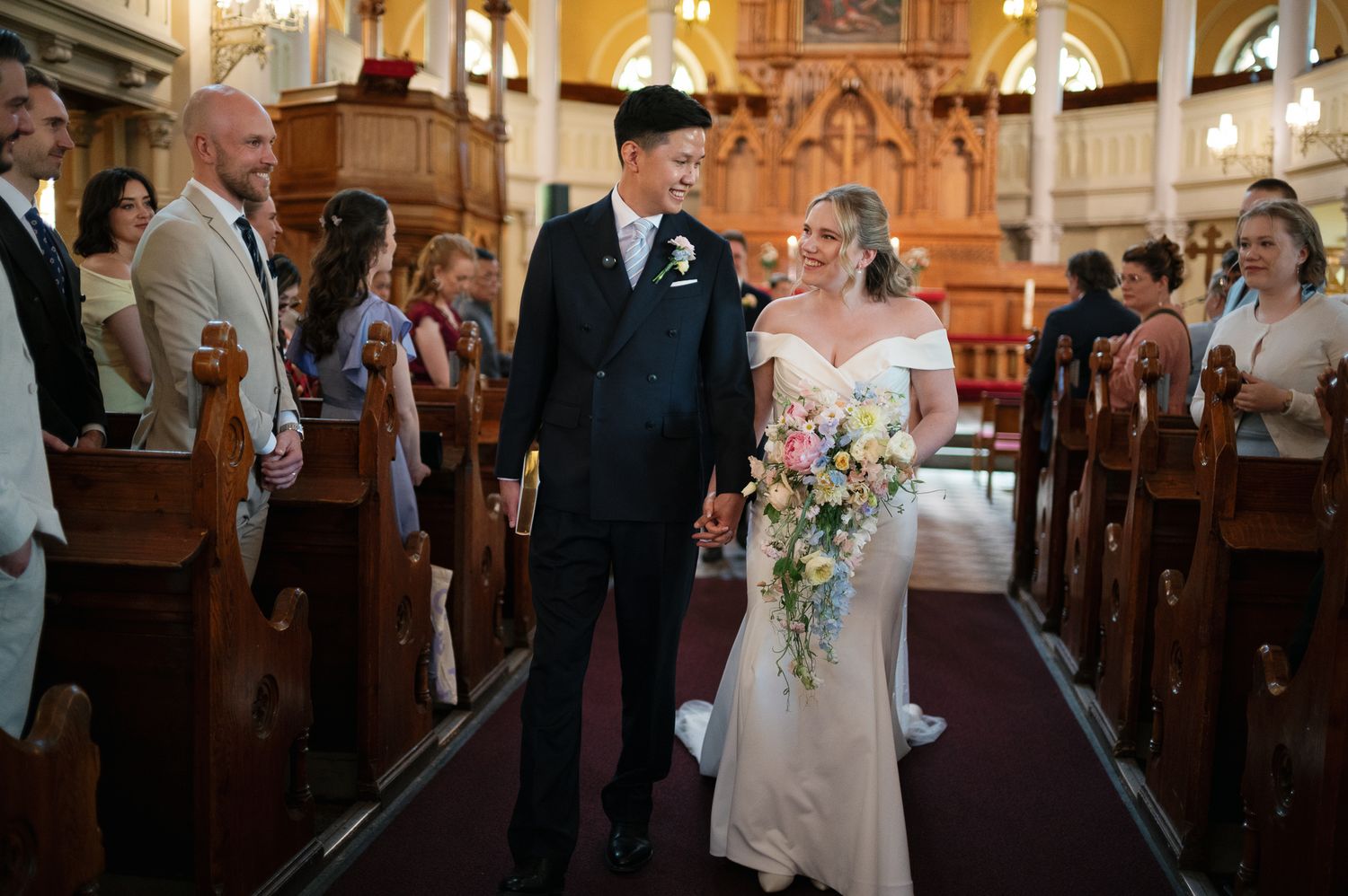 Bride and groom walk down church aisle holding hands, bride carries colorful cascading bouquet in elegant wedding ceremony.