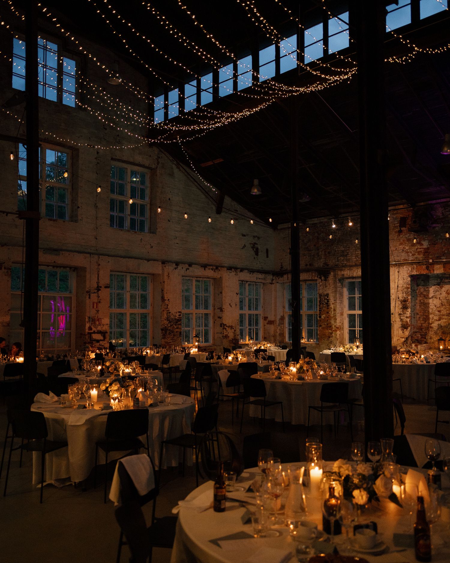 Romantic wedding reception in rustic industrial venue with candlelit tables and string lights overhead.