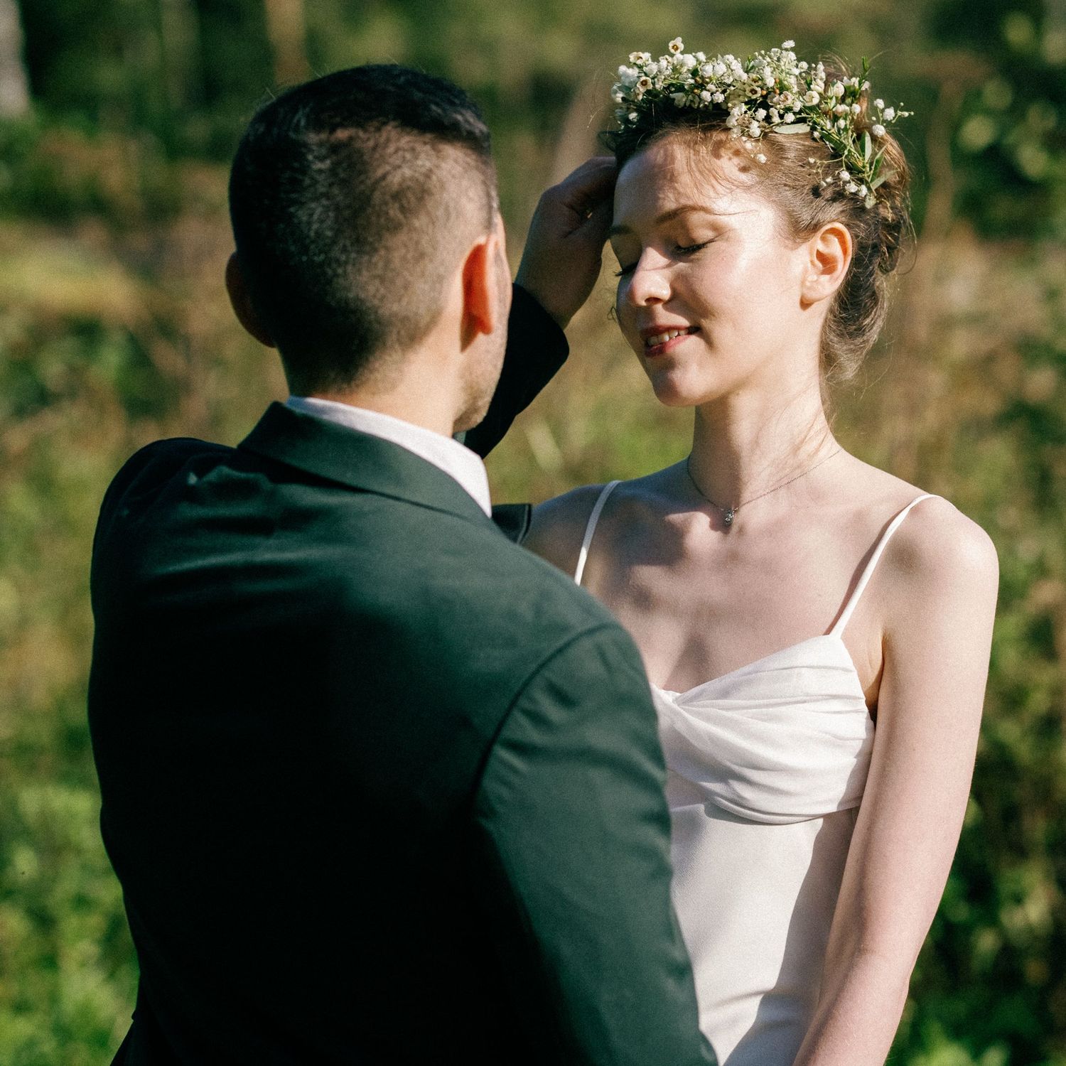 A tender moment between newlyweds outdoors, with the bride wearing a white gown and floral crown.