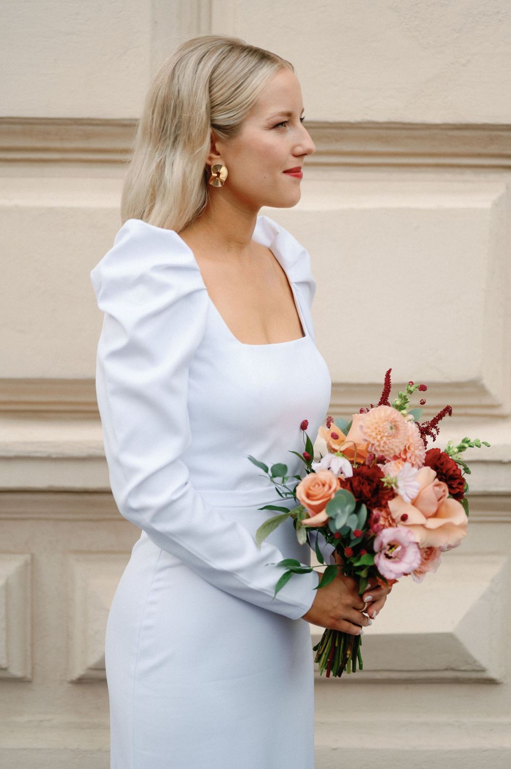 Bride in a modern white dress with puff sleeves holds a peach and coral floral bouquet against a light colored wall.