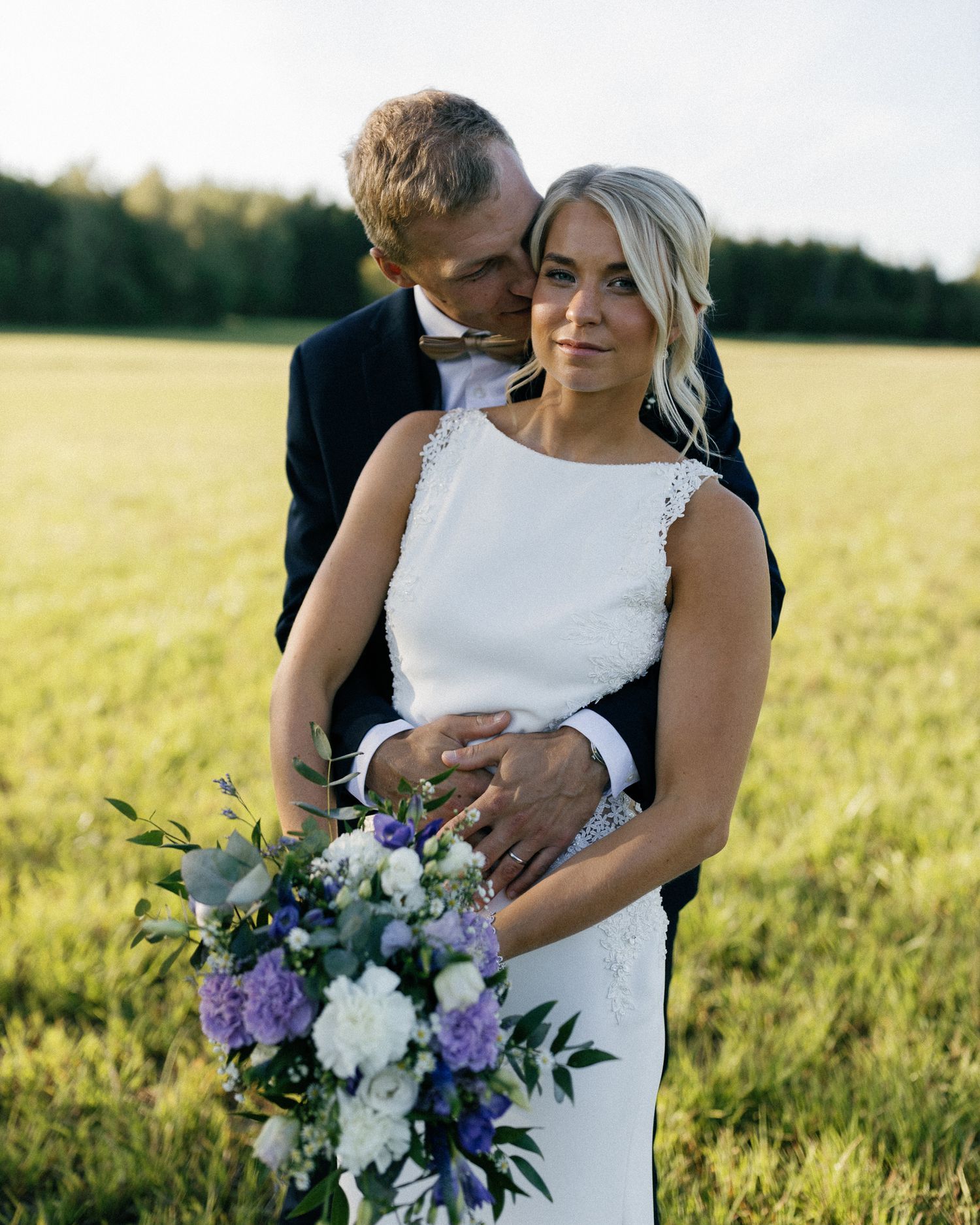 Bride in white dress holds purple and white bouquet while embracing groom in sunlit field.