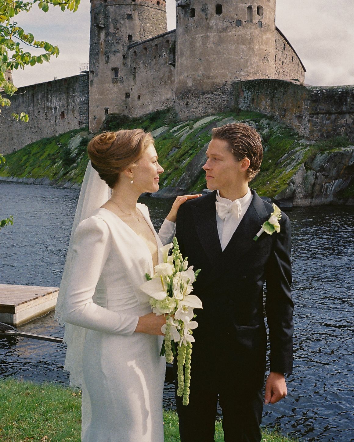 Bride and groom share a romantic moment by a lake with a historic stone castle in the background.