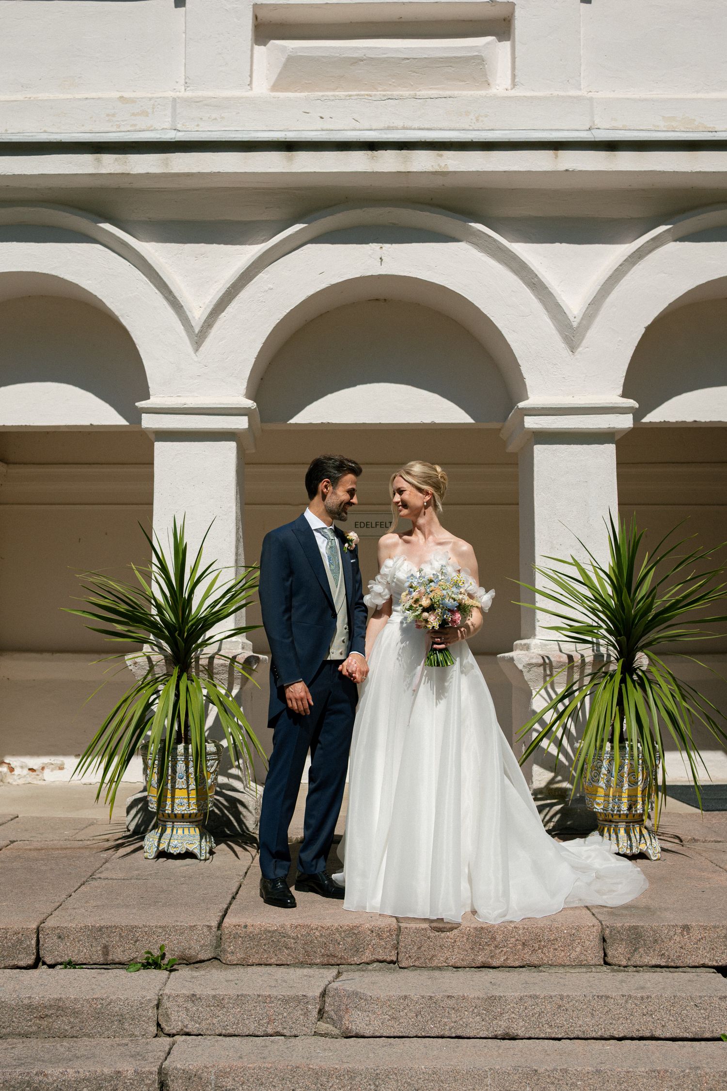 Bride and groom stand together before white arched building with tropical plants, bride holding bouquet in elegant white gown.