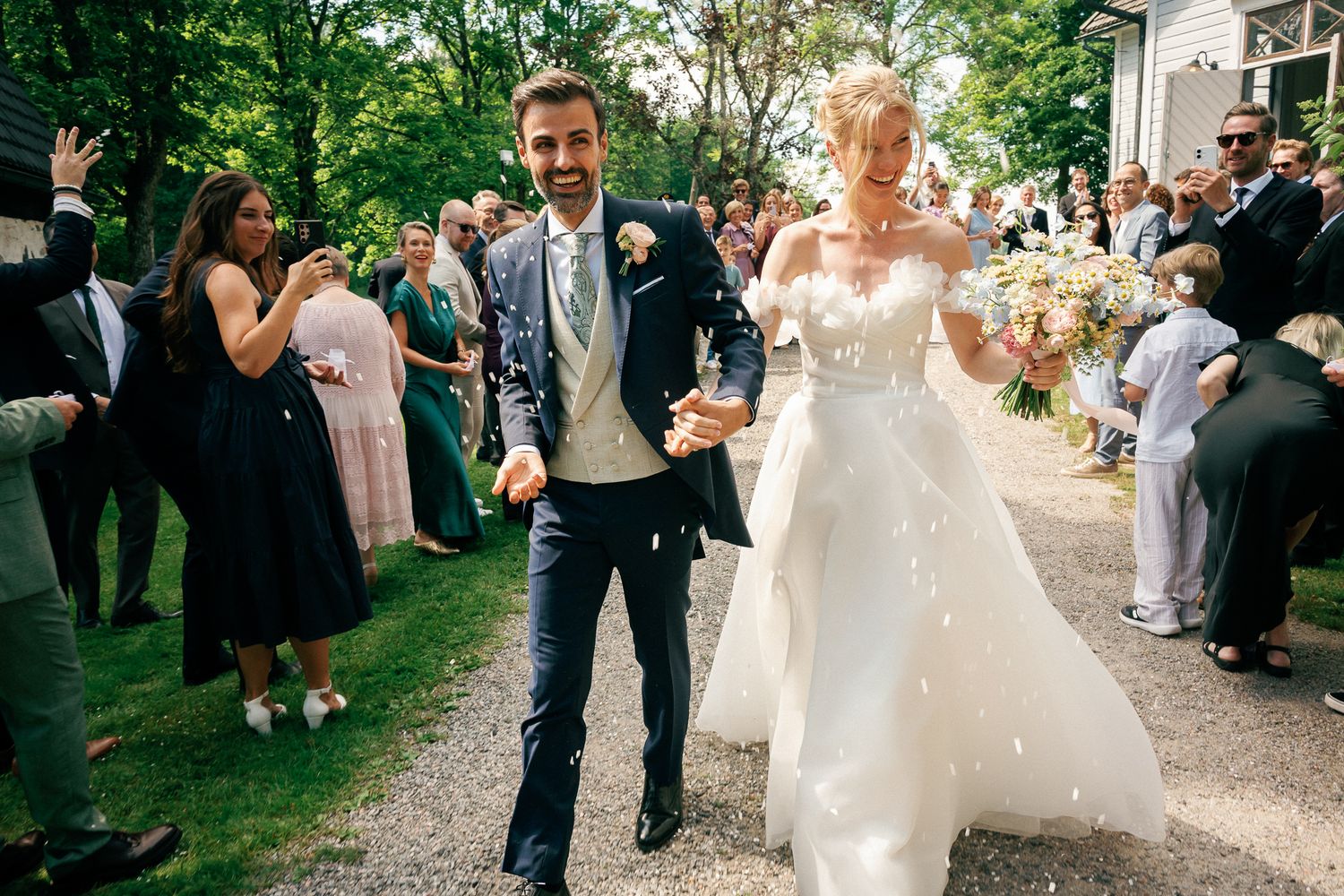 Bride and groom walk joyfully through confetti shower surrounded by cheering wedding guests outdoors.