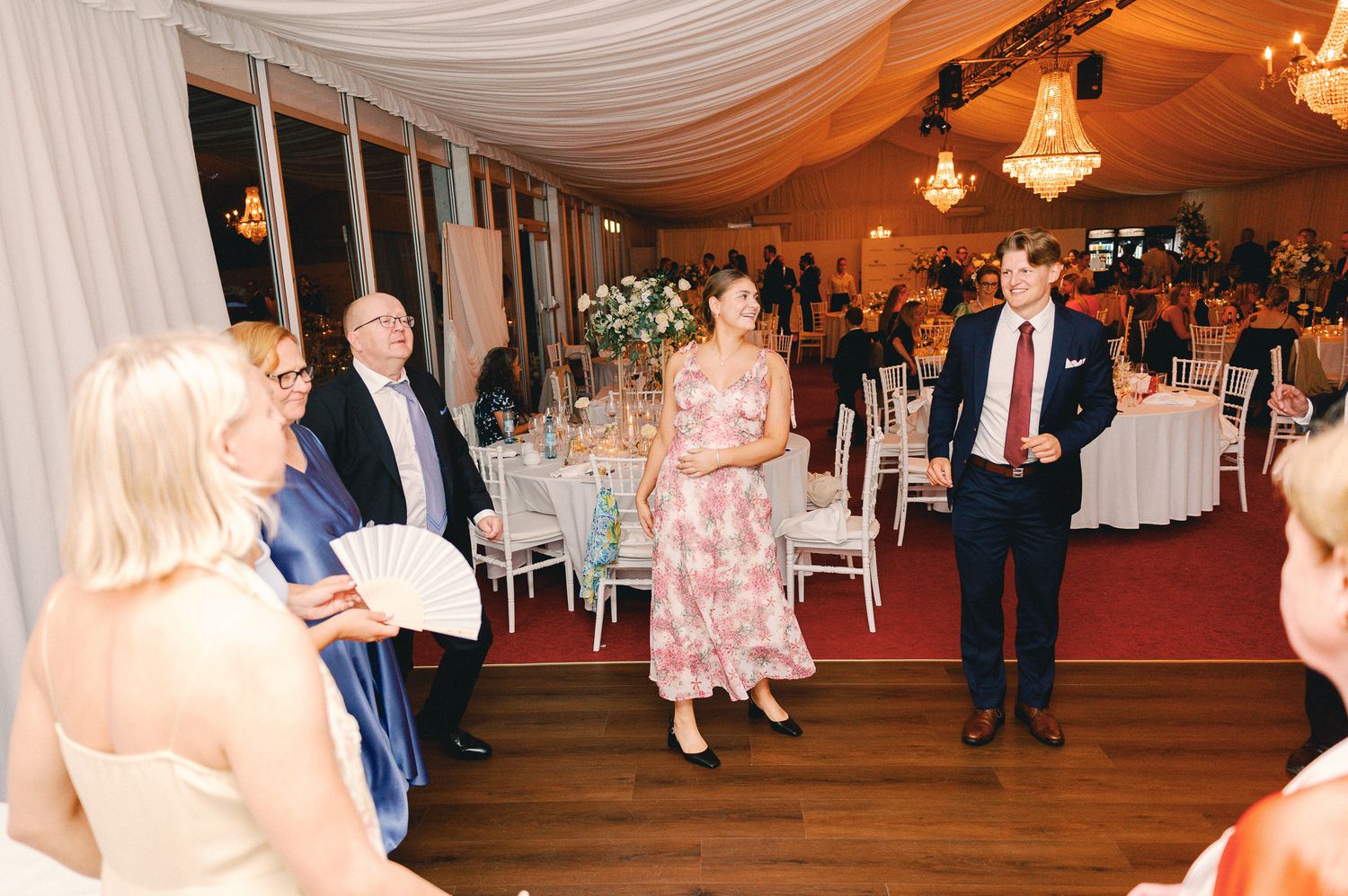 Elegant wedding reception in a marquee tent with guests mingling among floral centerpieces and chandelier lighting.