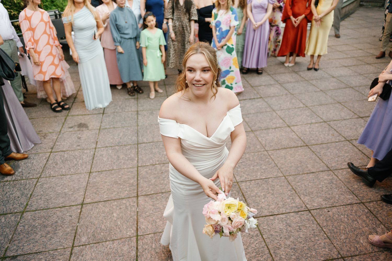 Bride in off-shoulder white gown smiles while holding pastel bouquet at outdoor wedding celebration.