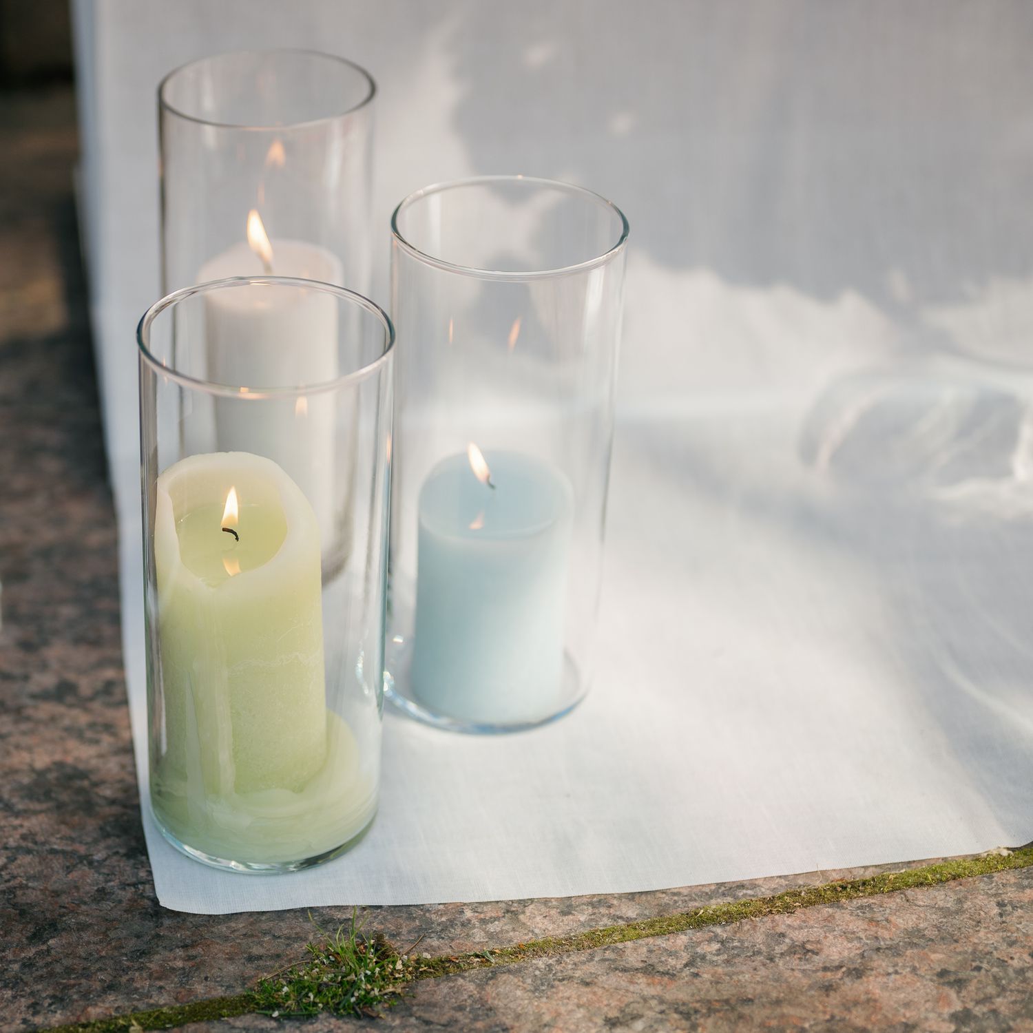 Three lit candles in tall glass cylinders, in green, white, and blue, arranged on a white cloth surface outdoors.