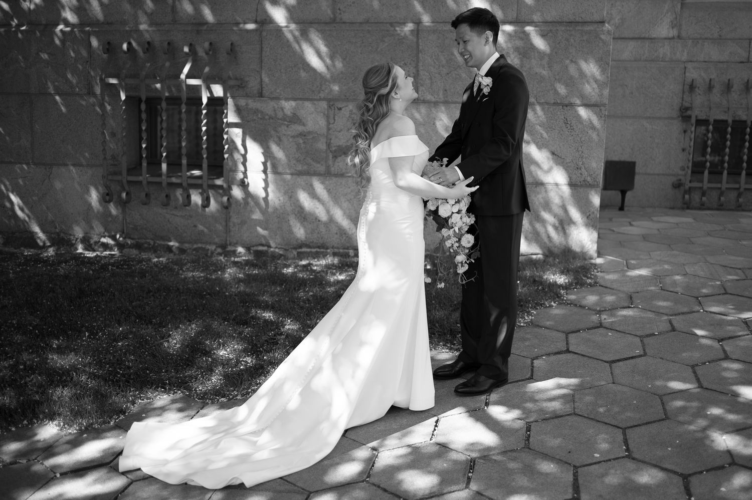 Black and white photo of bride in mermaid gown and groom holding hands outdoors on stone patio near stone wall.