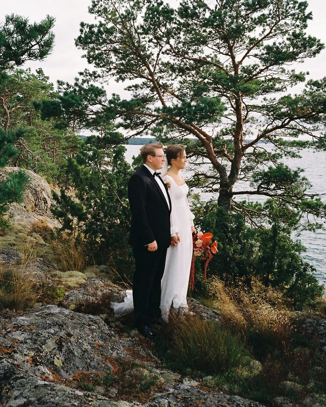 Wedding couple standing among pine trees on rocky coastal cliff.