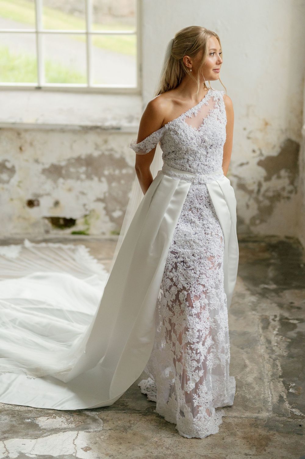 Bride in delicate lace gown with dramatic train standing in sunlit rustic interior.