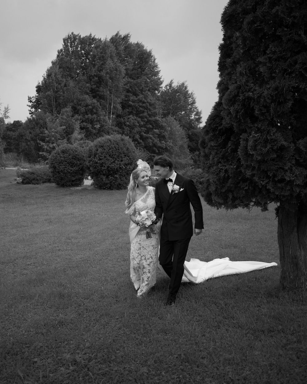 A wedding couple walks hand in hand through a scenic field with tall trees in black and white.