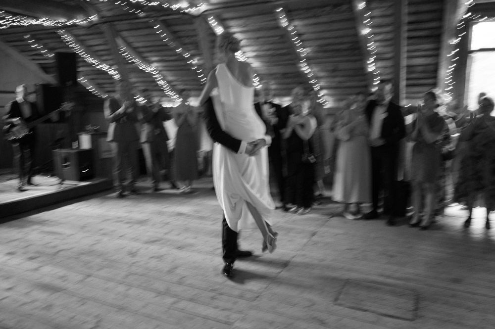 Motion-blurred black and white photo of dancing at a wedding reception in a rustic venue.