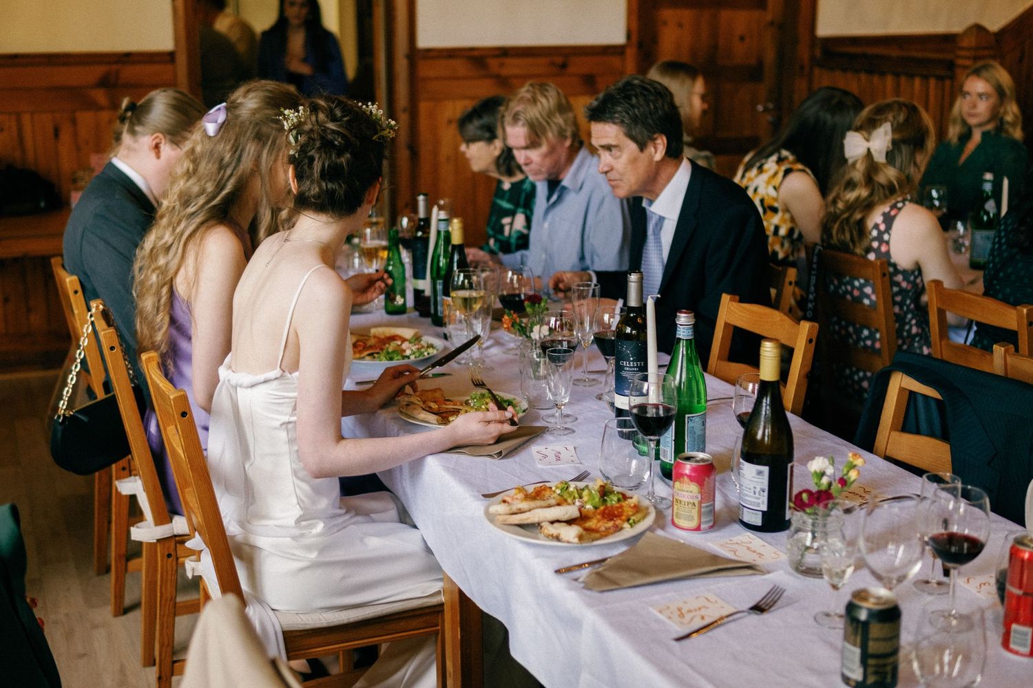 Wedding celebration with guests dining at rustic wooden tables decorated with wine bottles and glassware.