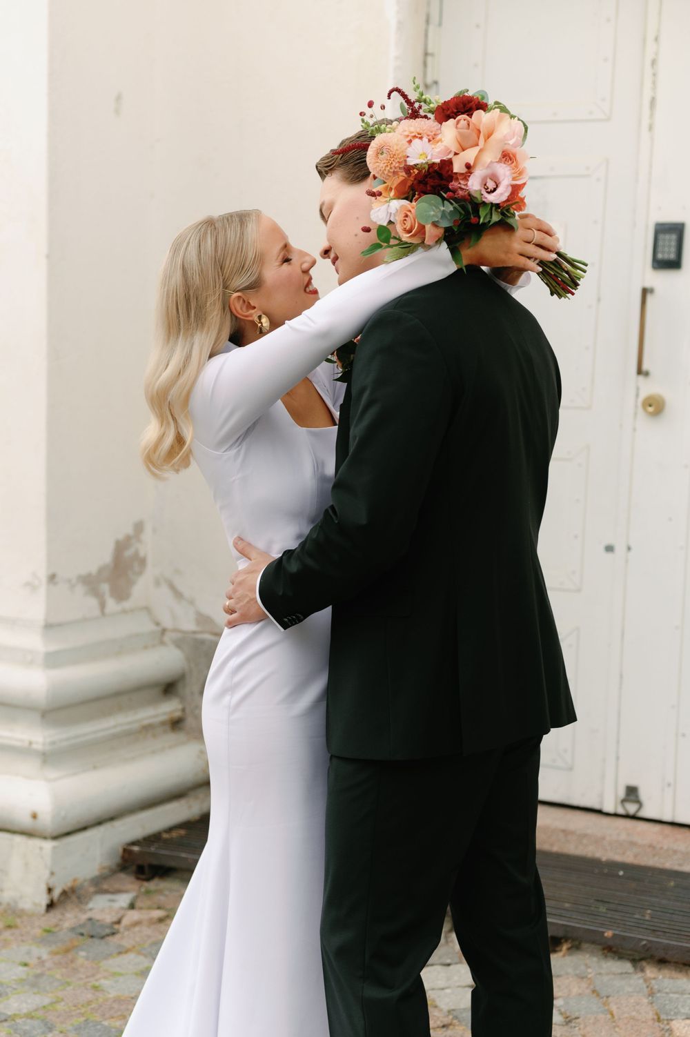 A tender wedding moment with the bride in white and groom in black sharing an embrace near a white door.