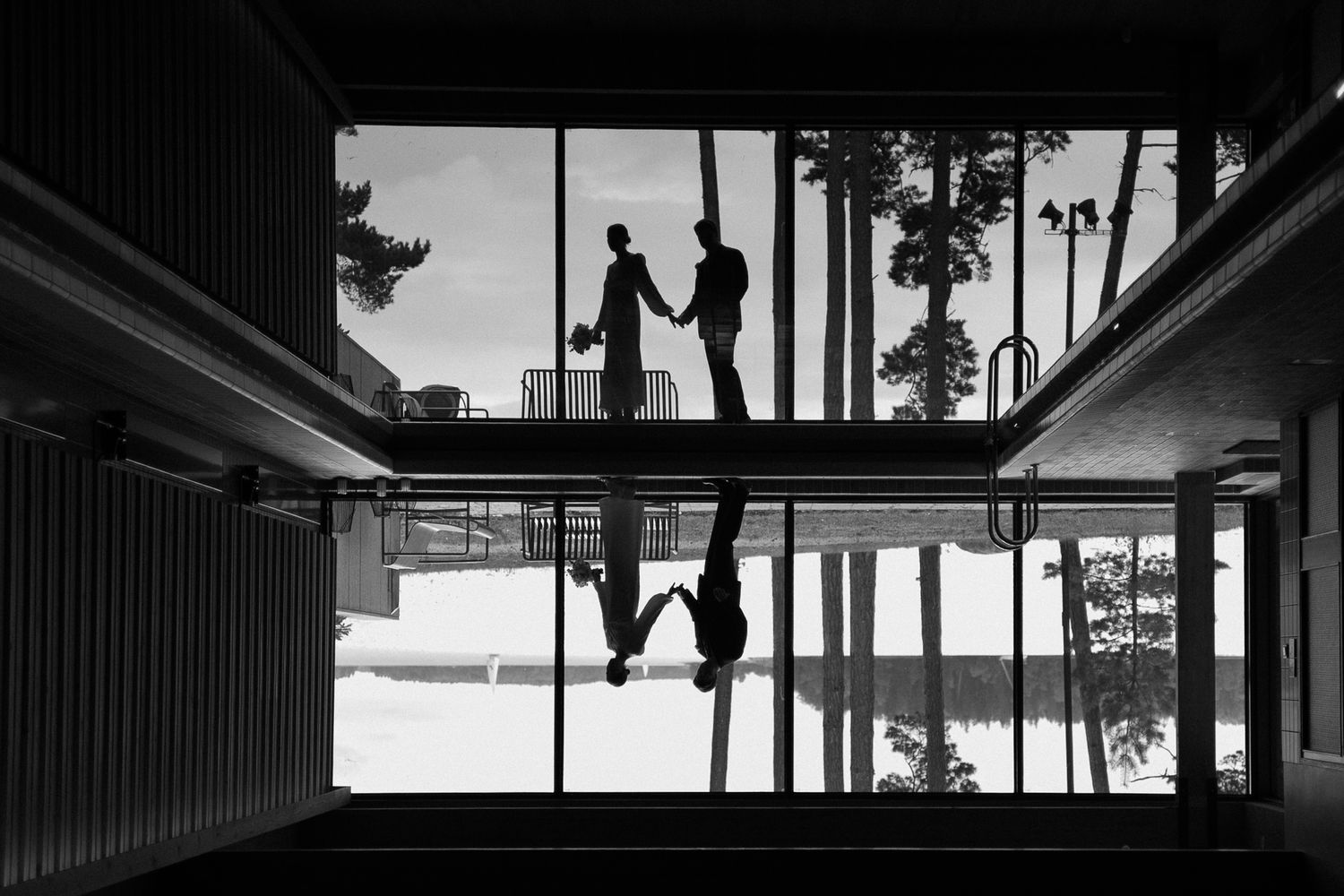 Black and white silhouette of two people shaking hands reflected in glass building facade with tall trees backdrop.