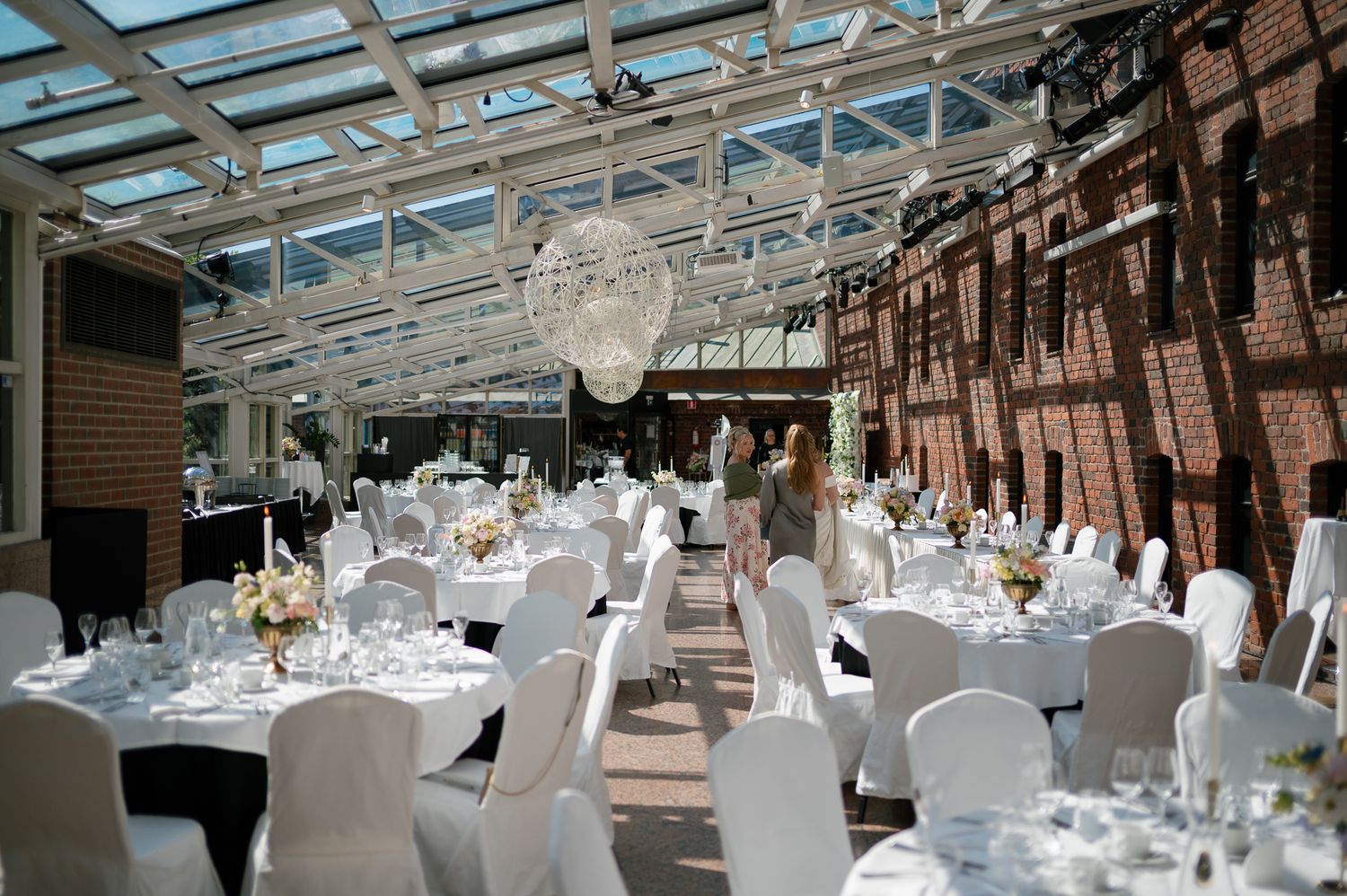 Elegant wedding reception venue with white chair covers, round tables, crystal chandelier, and a stunning glass ceiling atrium.