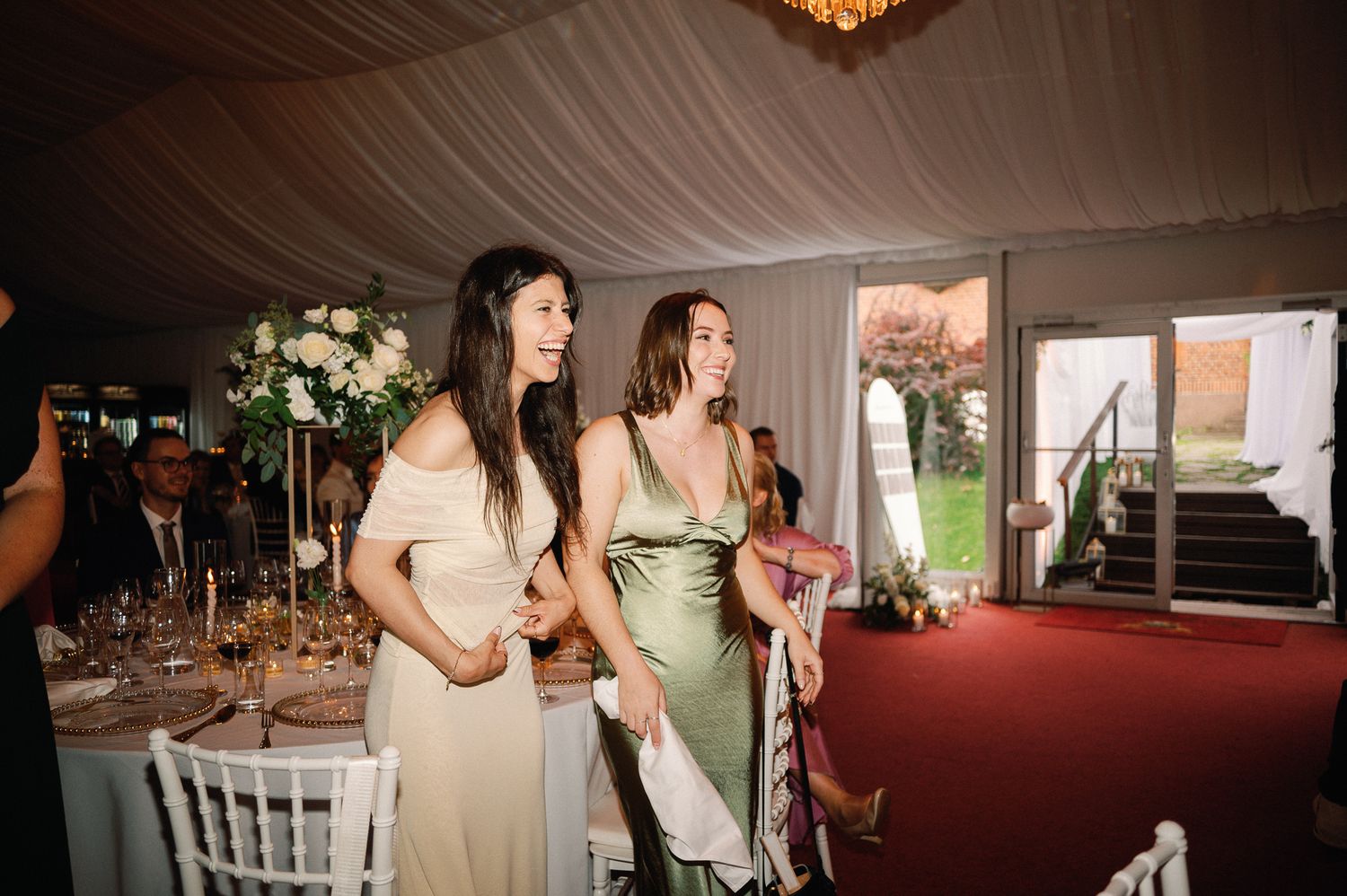 Two bridesmaids in elegant gowns smiling and walking into a beautifully decorated wedding reception tent.