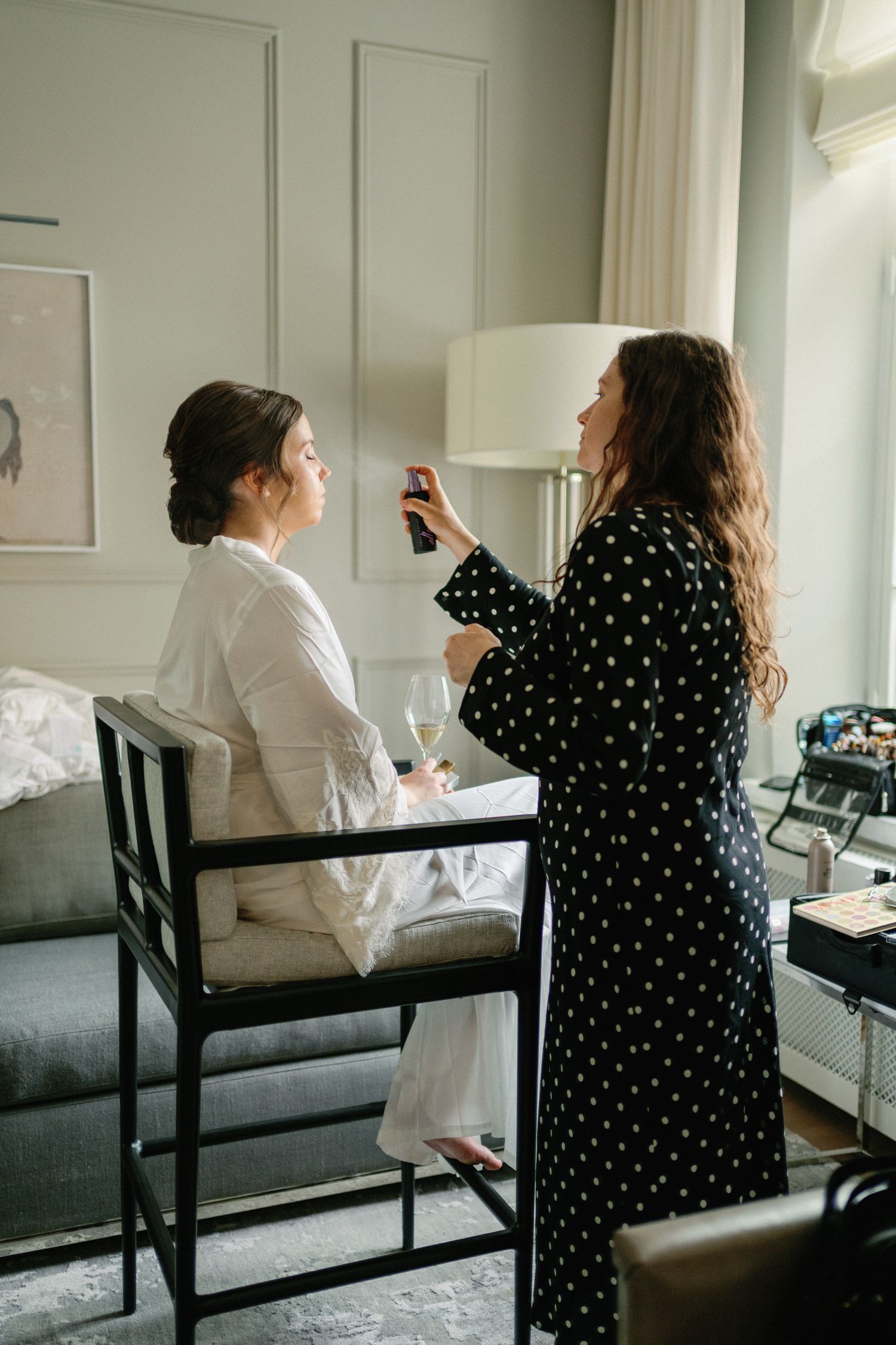 Makeup artist in polka dot robe applying finishing spray to bride in white robe seated in elegant hotel room.
