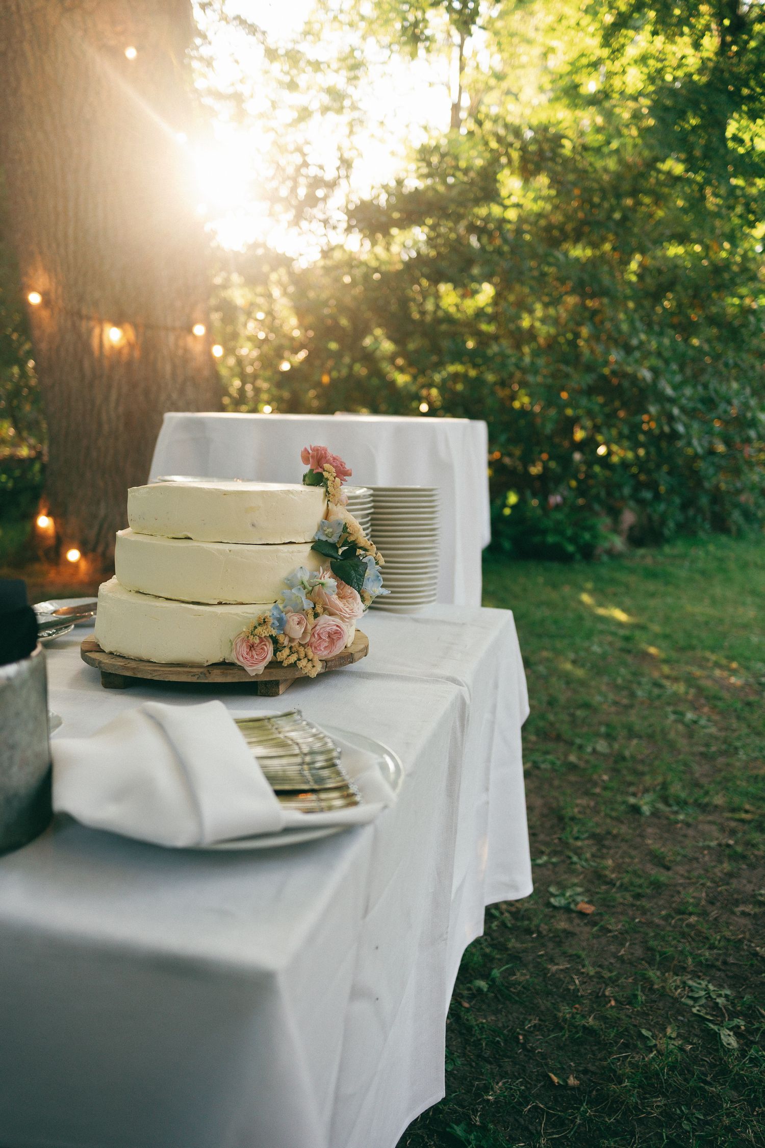 Three-tier white wedding cake with pink flowers on outdoor table, golden sunlight filtering through trees at garden reception.