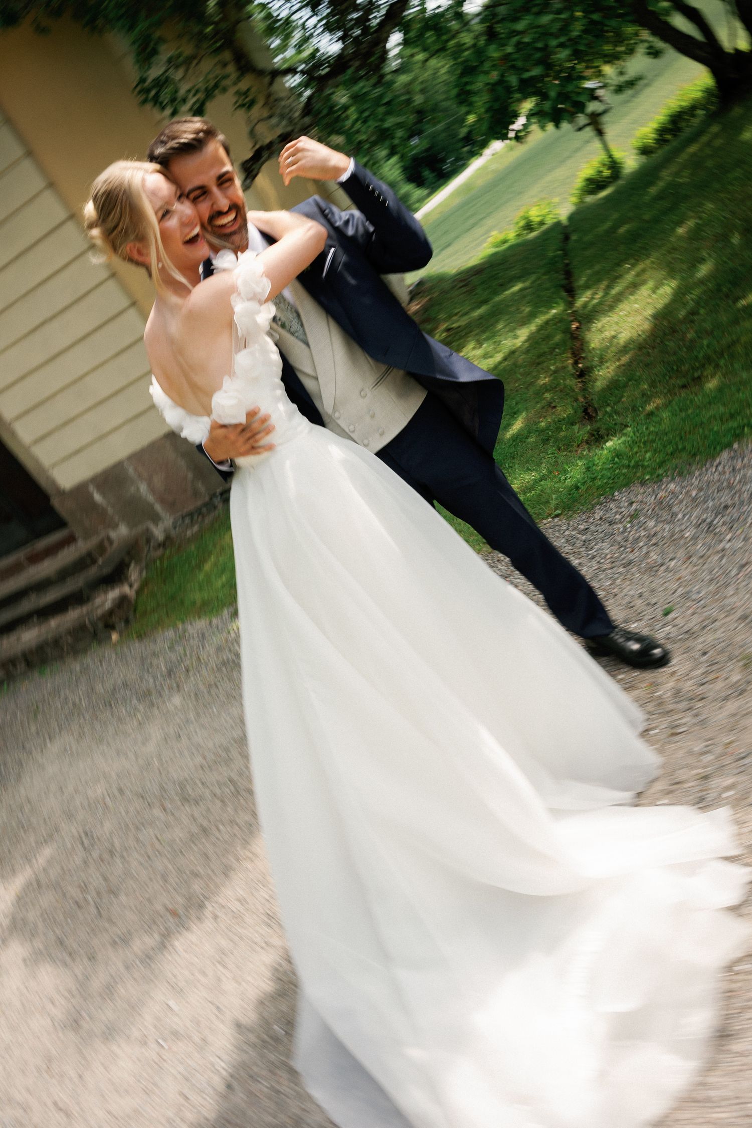 A joyful groom in a navy suit twirls his smiling bride in a white flowing gown outdoors on their wedding day.