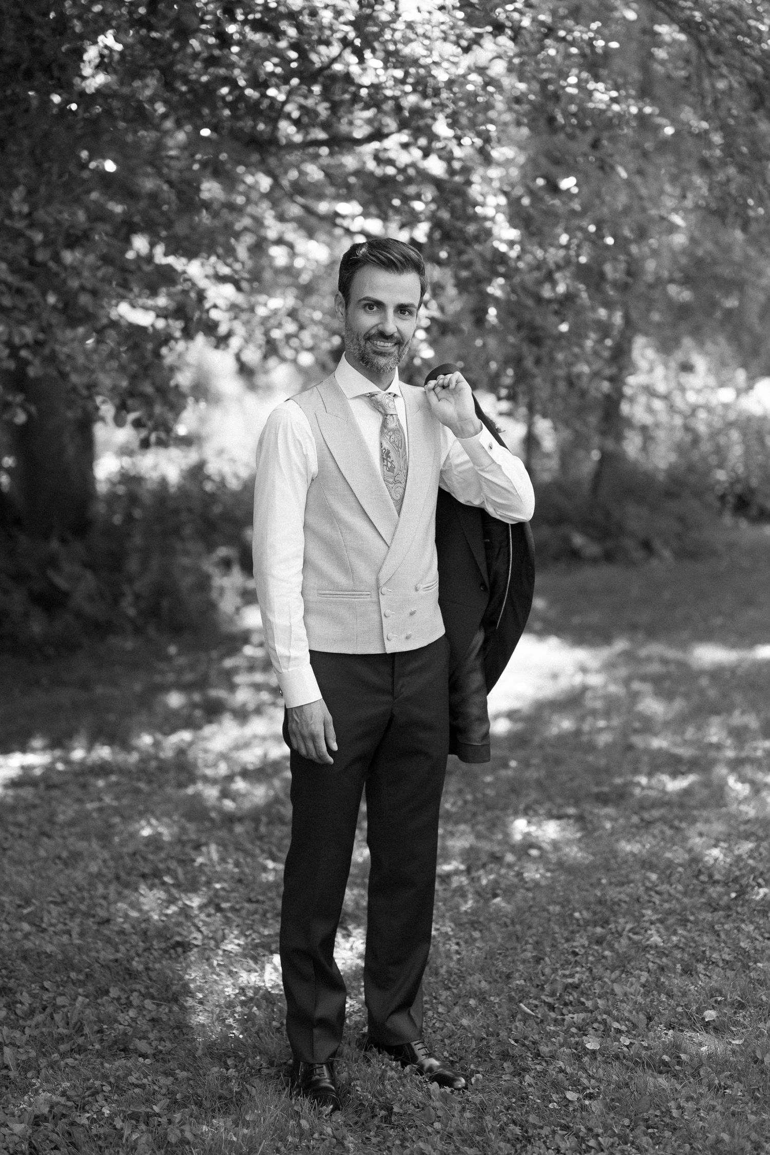 A smiling groom in a white vest and tie holds his jacket over his shoulder in a black and white outdoor portrait.