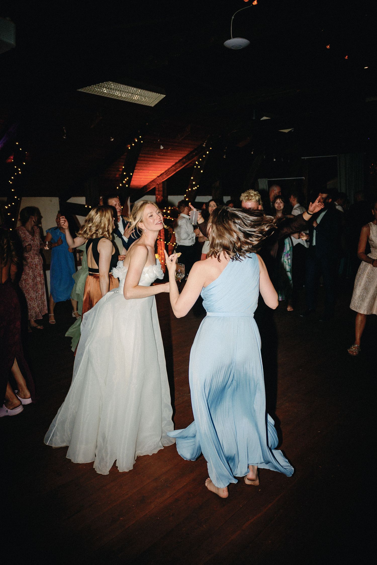 Bride in white gown dancing with bridesmaid in light blue dress at a lively wedding reception.
