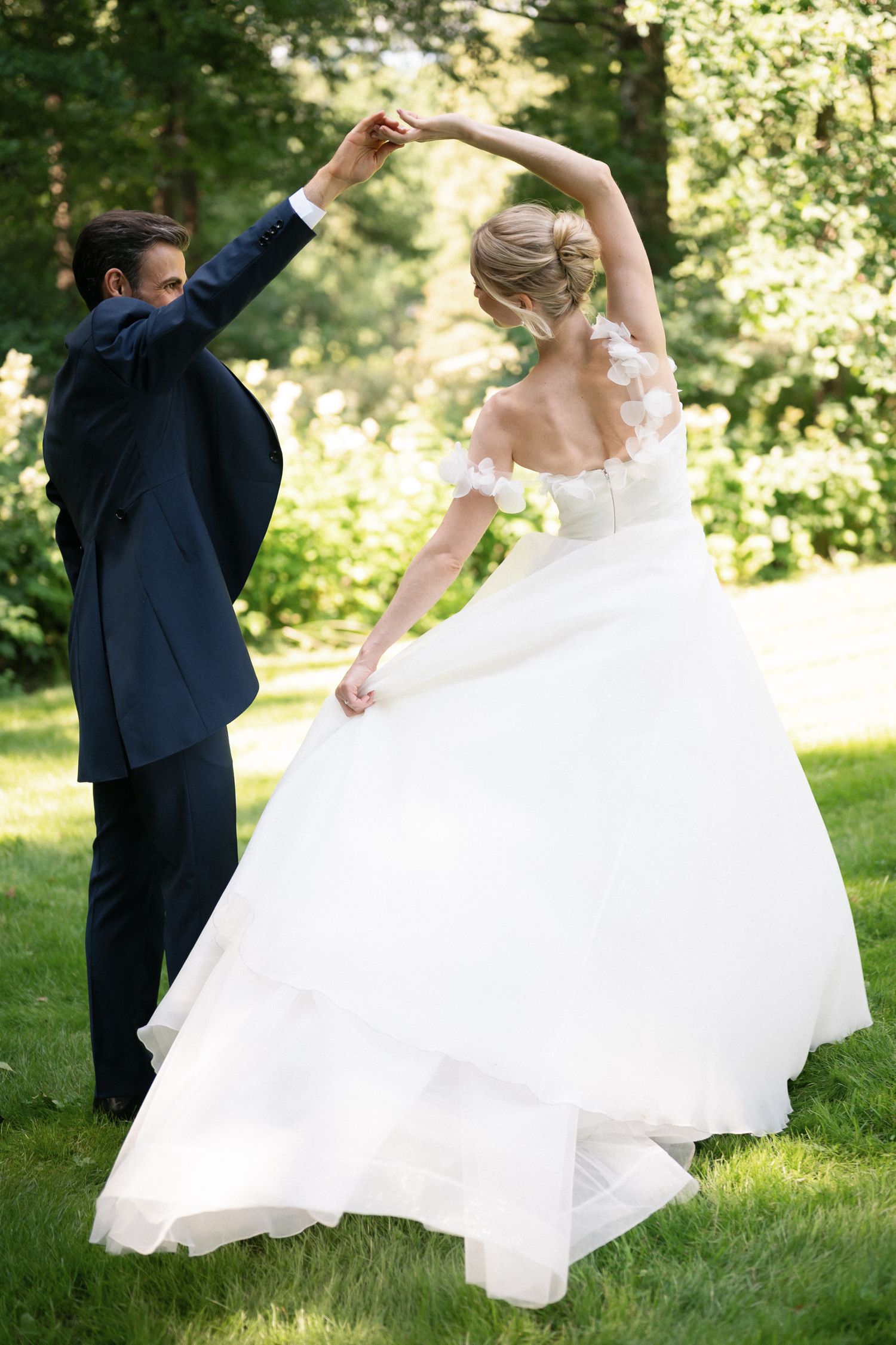 Bride in white ballgown twirls as groom in navy suit holds her hand in a sunlit green garden.