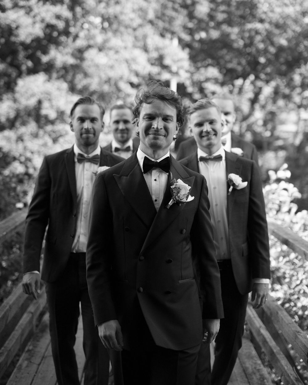 Groomsmen in black tuxedos standing together on wooden deck outdoors.
