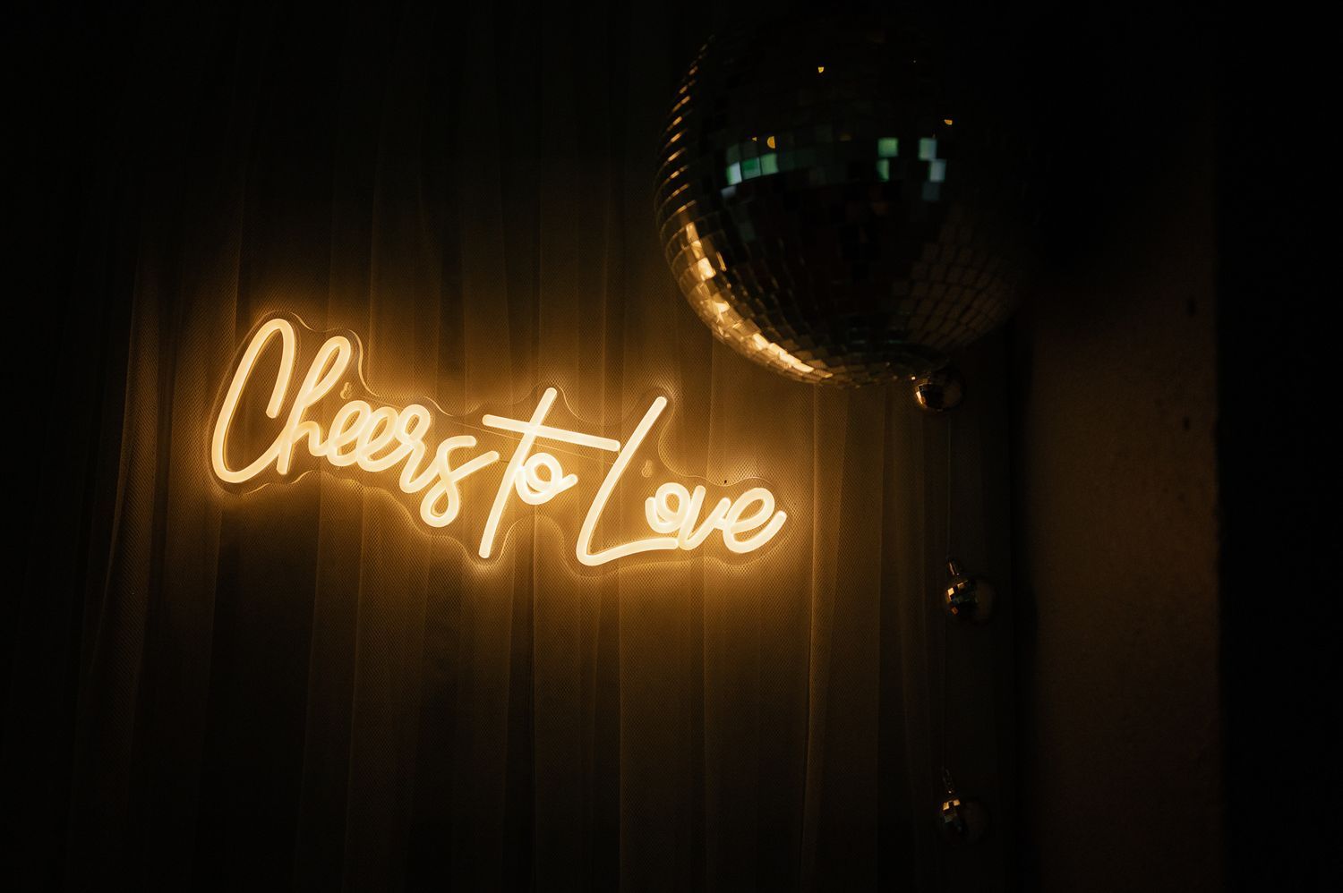 Neon 'Cheers to Love' sign glows beside a disco ball in dark ambiance.