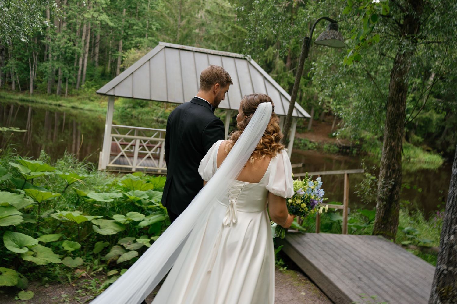 A couple walks along a garden path with flowing white dress near a wooden gazebo.