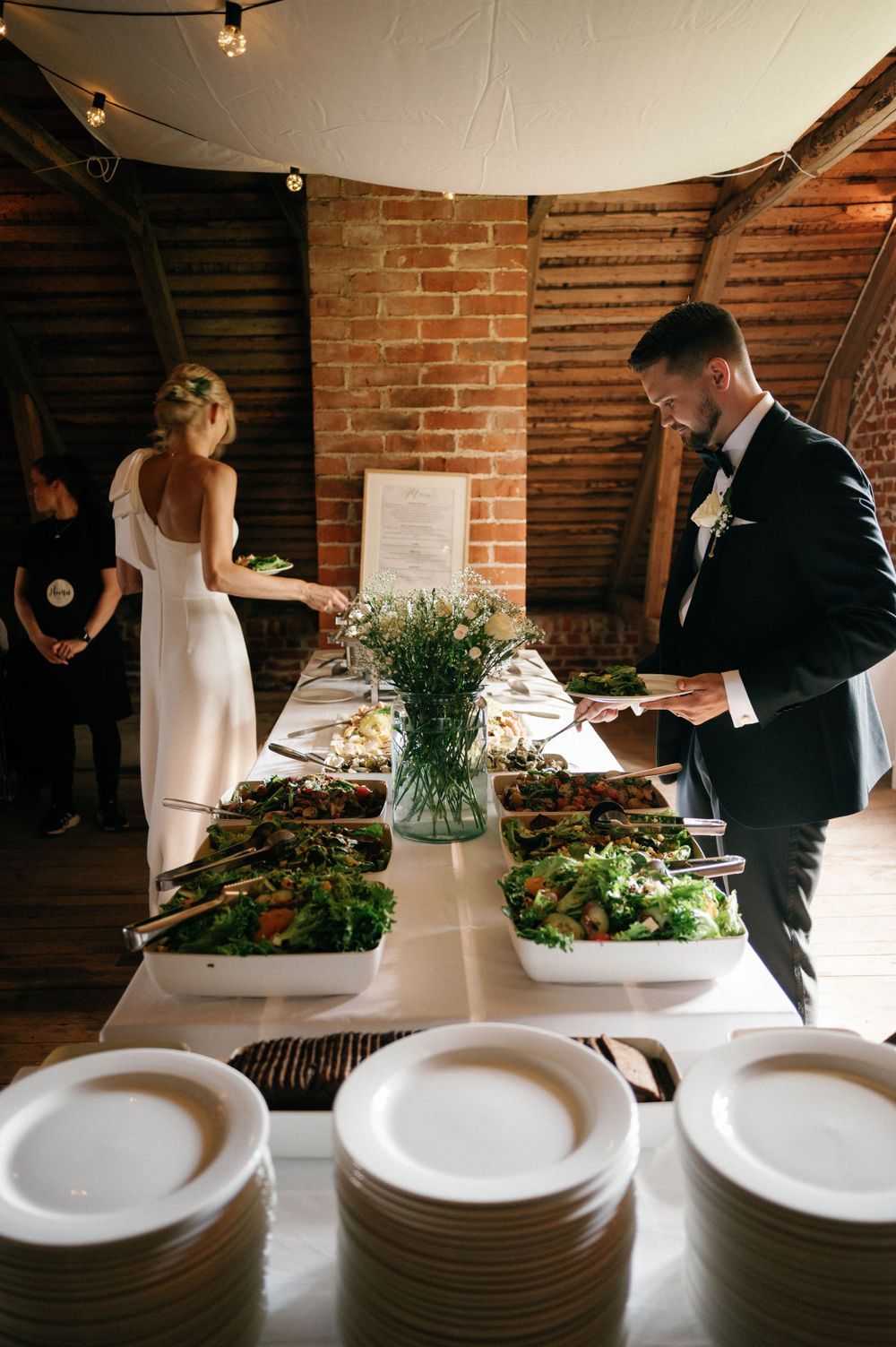 Elegant buffet setup with white plates and fresh greenery against exposed brick walls.