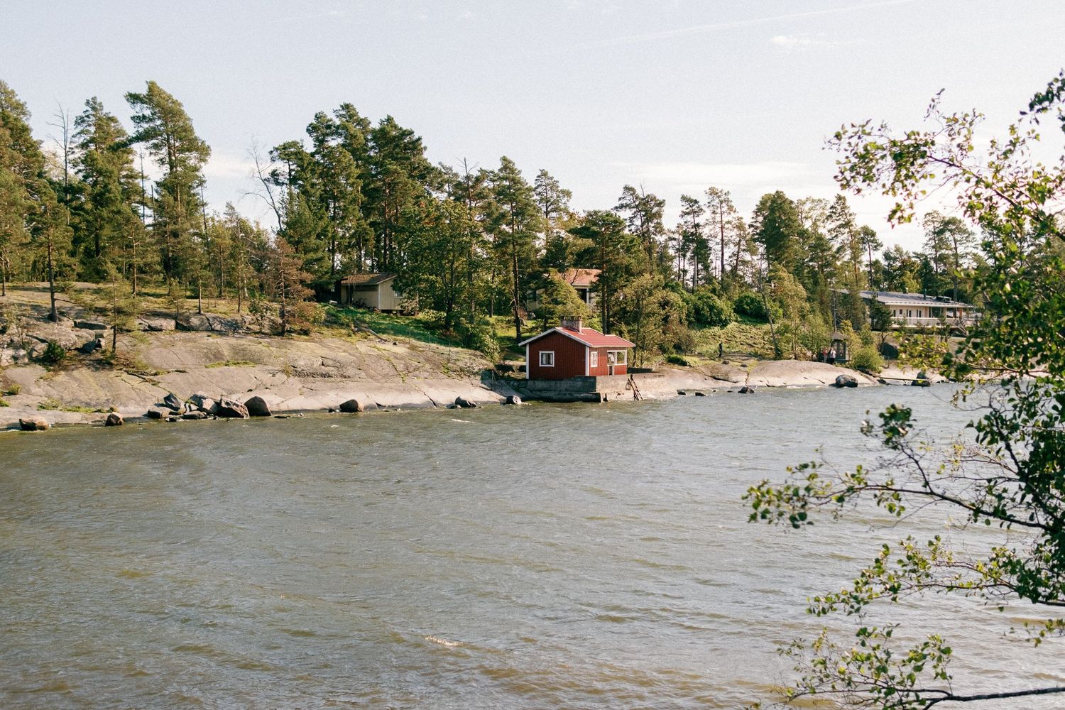 Red cottage on rocky coastline surrounded by trees and water.
