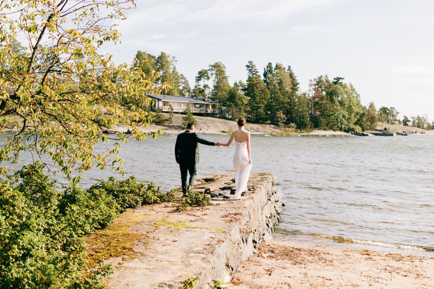 A couple walks hand in hand along a rocky shoreline with evergreen trees in the background.