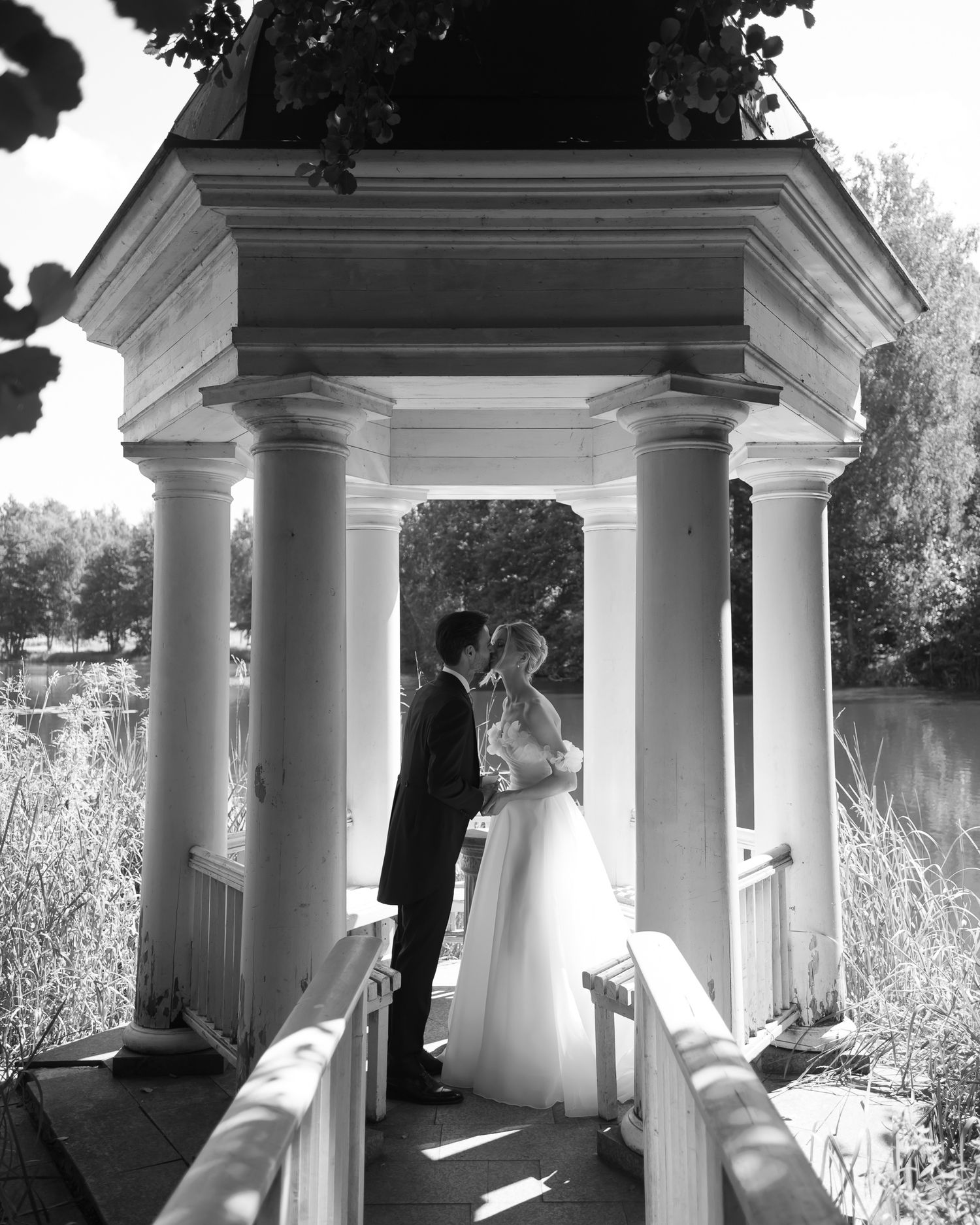 Black and white photo of a bride and groom kissing under an elegant gazebo with columns near a lake.