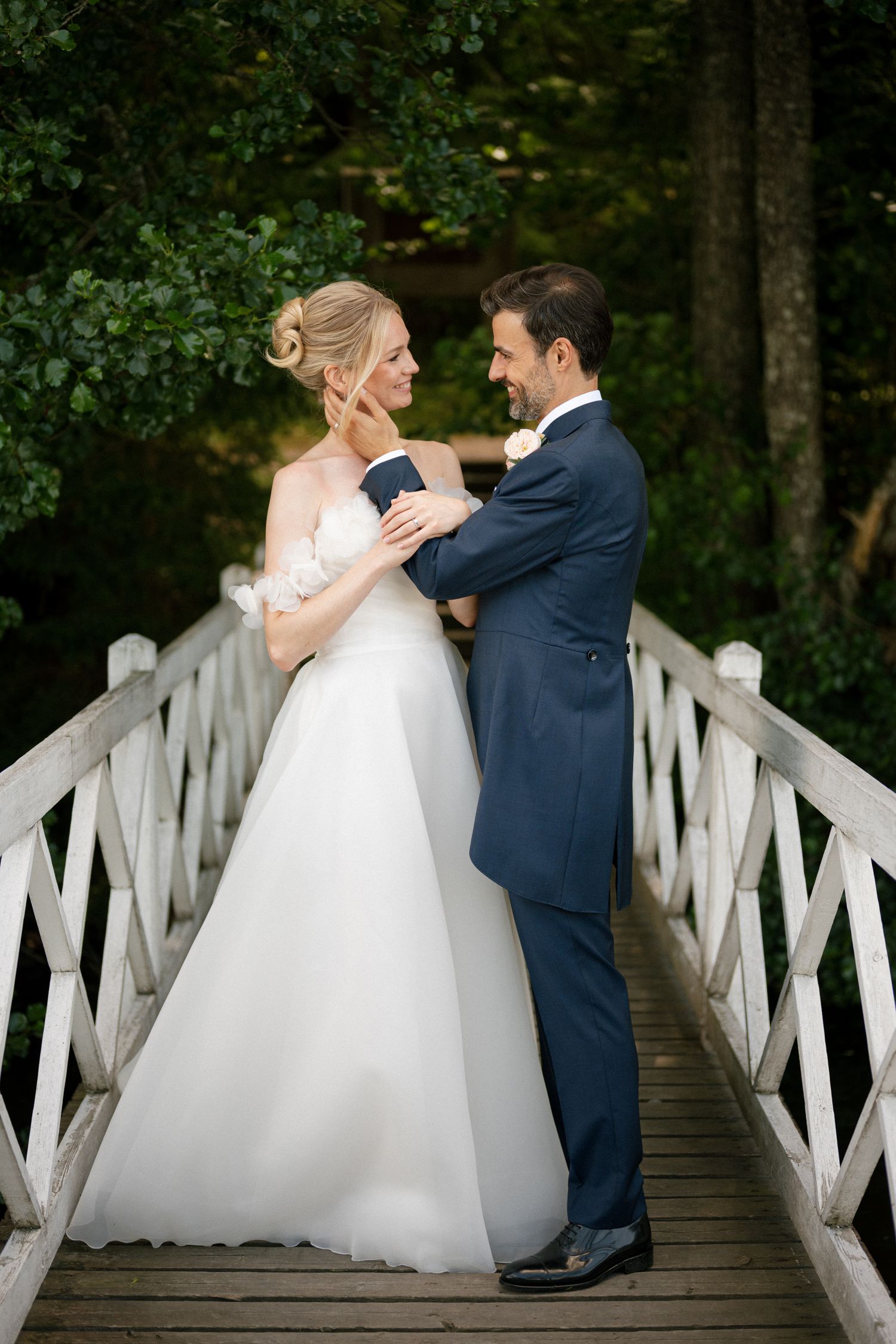 Bride and groom share a romantic moment on a white wooden bridge surrounded by lush green trees.