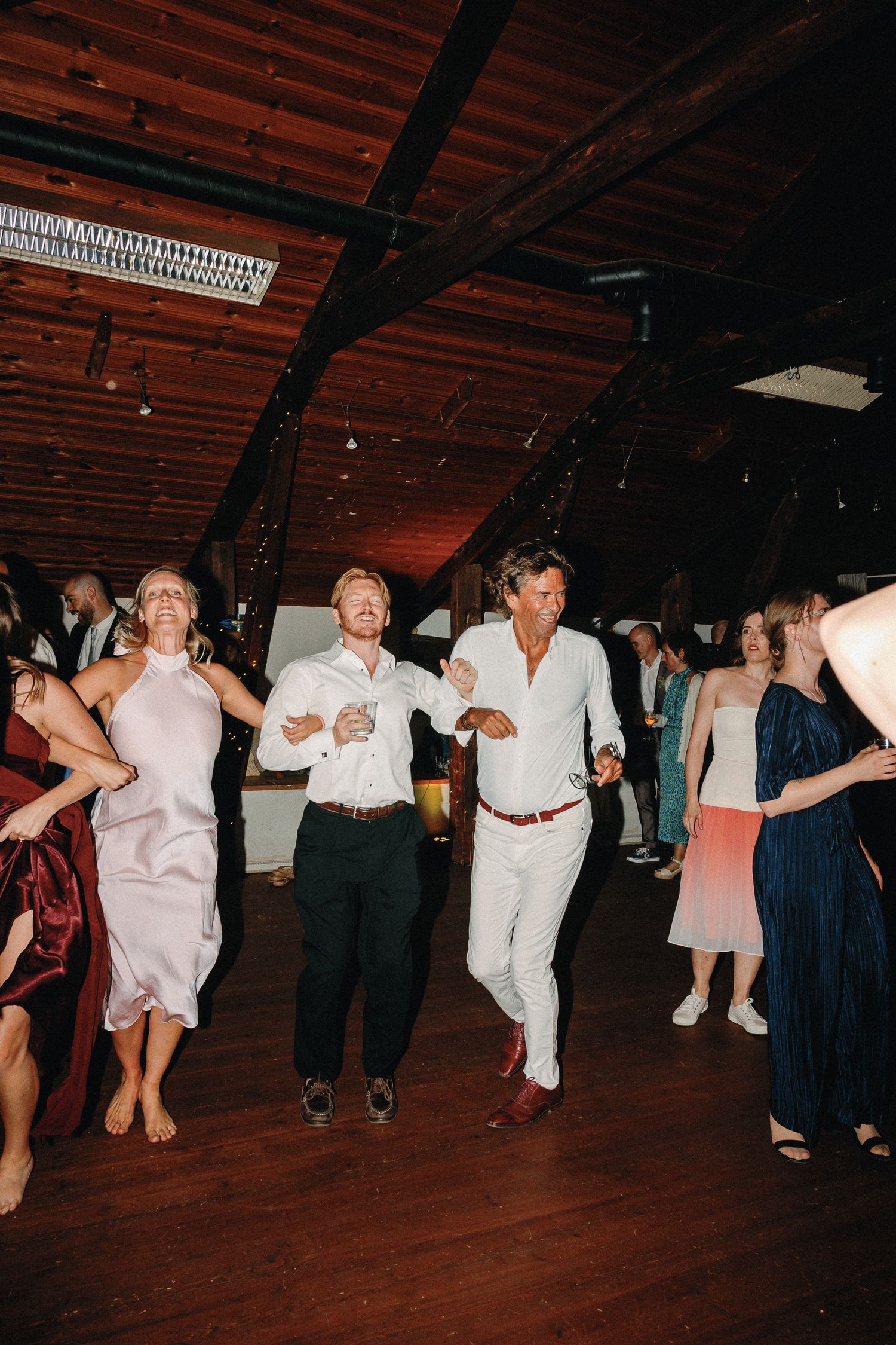 Guests dancing joyfully at an indoor wedding reception with wooden ceiling and warm ambient lighting.