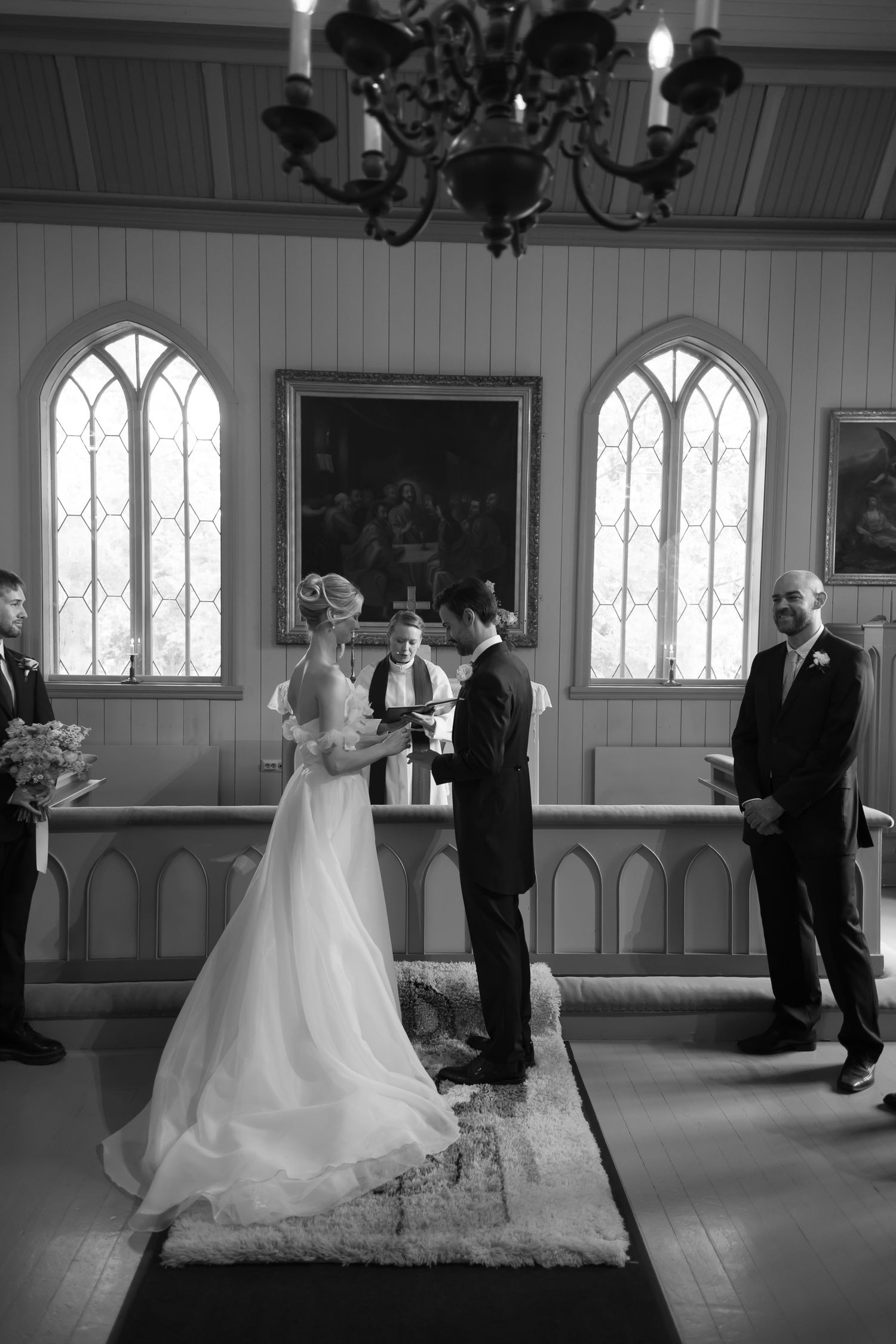 Black and white photo of a bride and groom exchanging vows at a church altar with gothic windows and a chandelier.