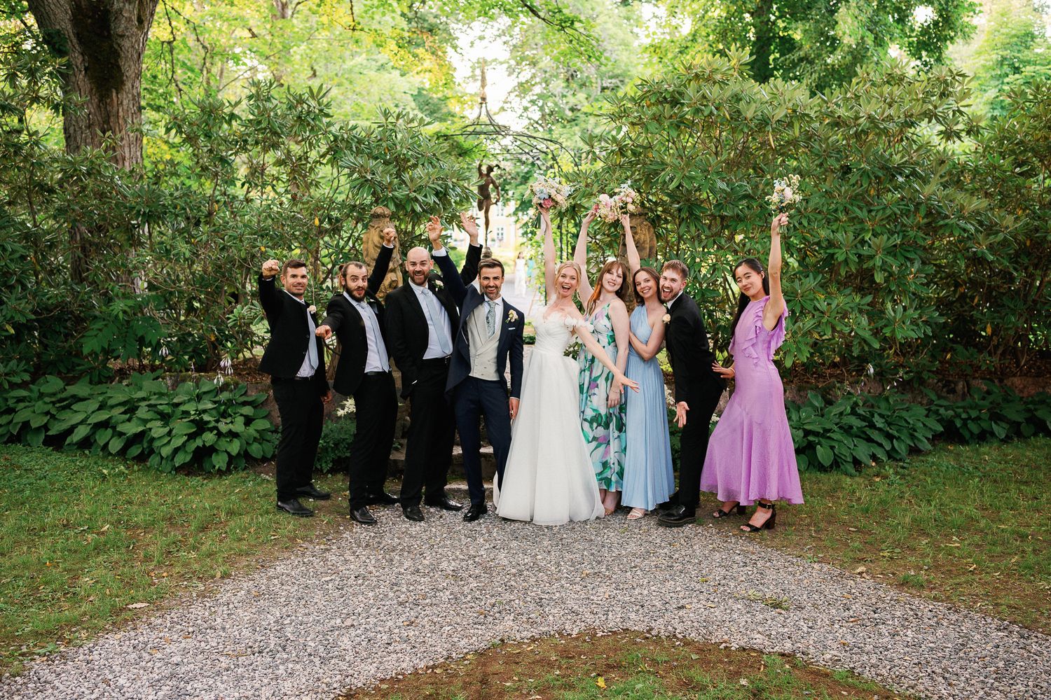 Wedding party celebrating outdoors on gravel path surrounded by lush green trees, bride in white gown with joyful guests.