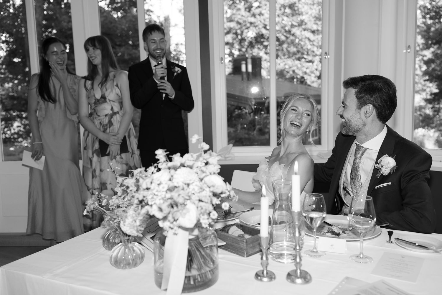 Black and white wedding photo of bride and groom at table laughing with guests giving speech in background.