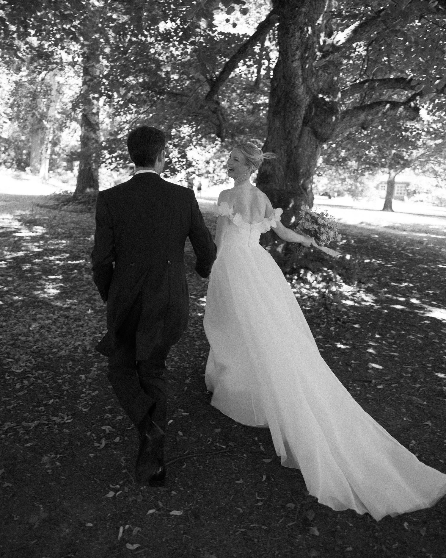 Black and white photo of a bride in a flowing gown and groom in a suit walking hand in hand under trees.