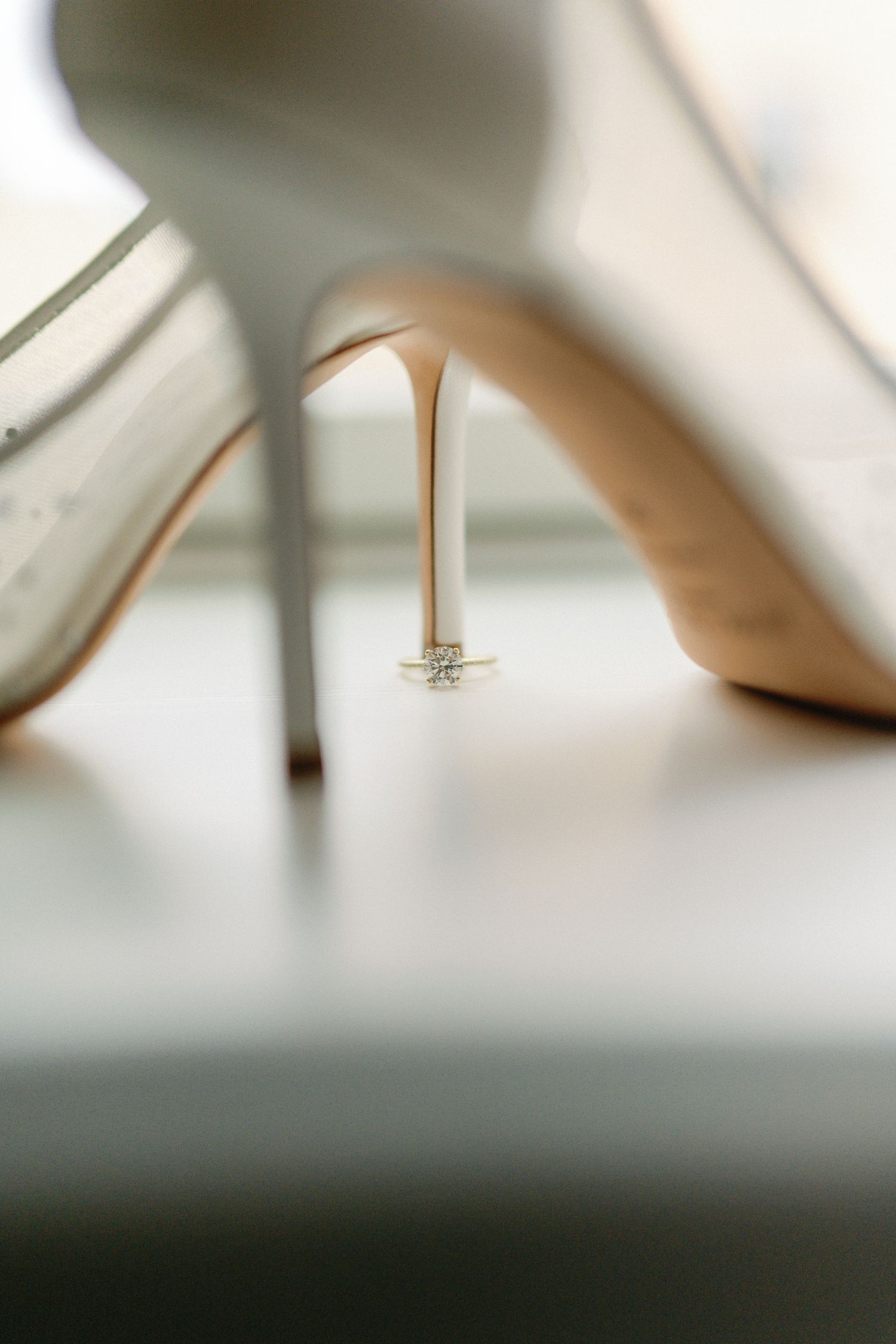 White bridal heels with a diamond engagement ring resting beneath the stiletto heel on a white surface.