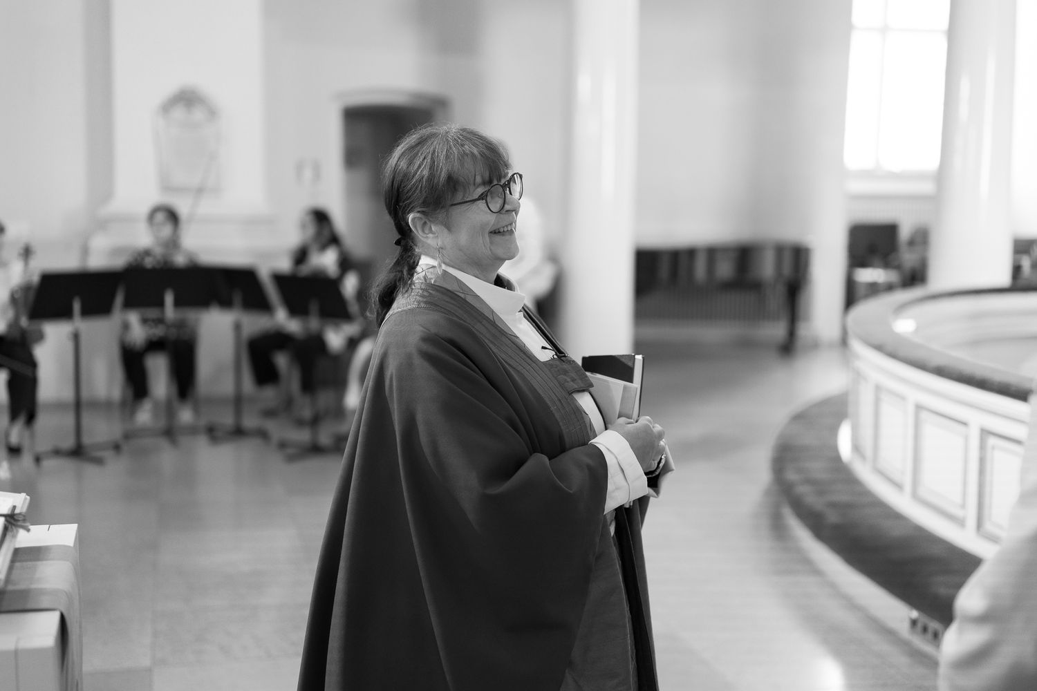 Black and white photo of a person in a cape standing in a church hall with musicians in the background.