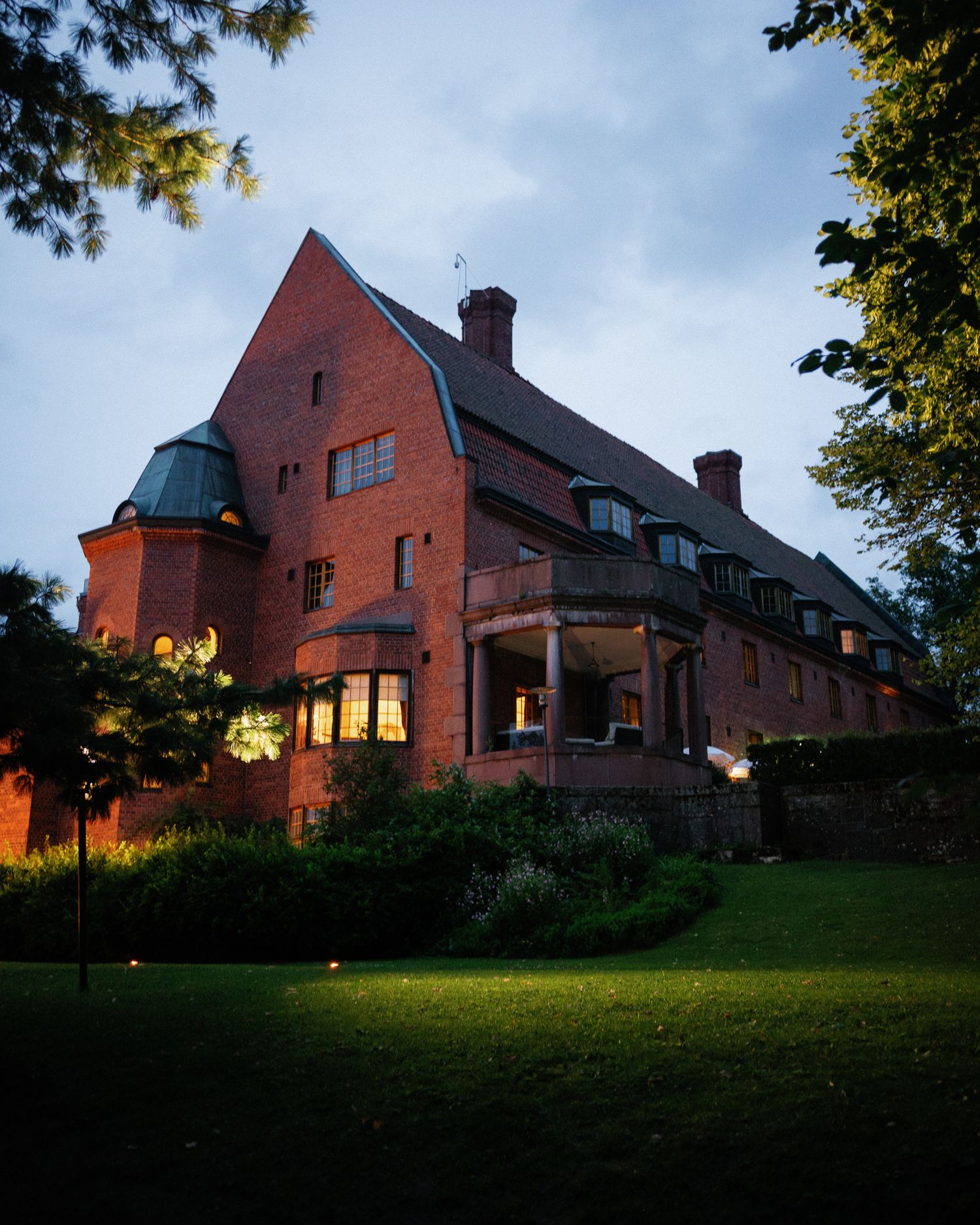 A grand red brick manor house photographed at dusk with warm glowing windows surrounded by lush green lawns and trees.