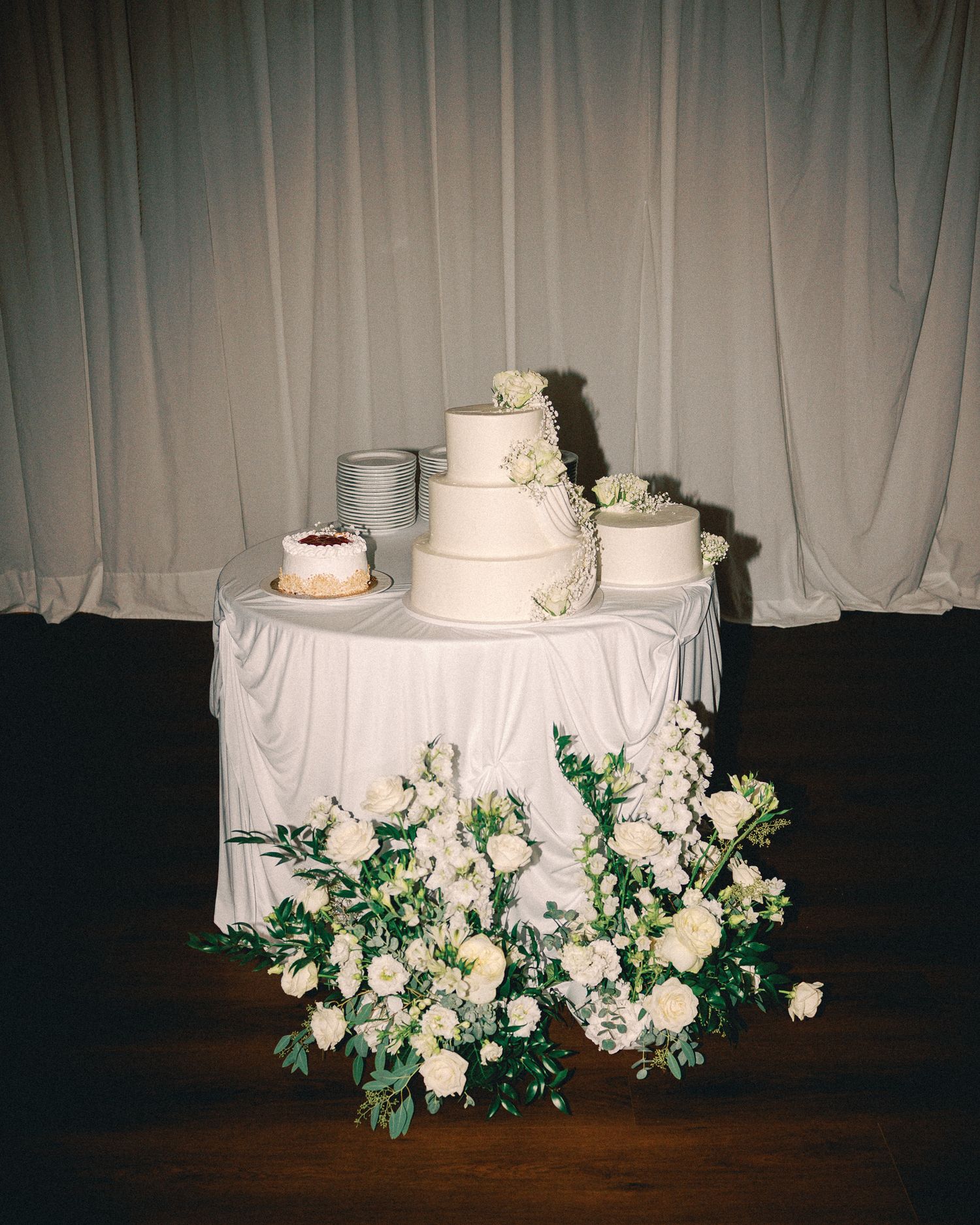 Elegant three-tier white wedding cake on draped table with lush white floral arrangements and greenery at base.