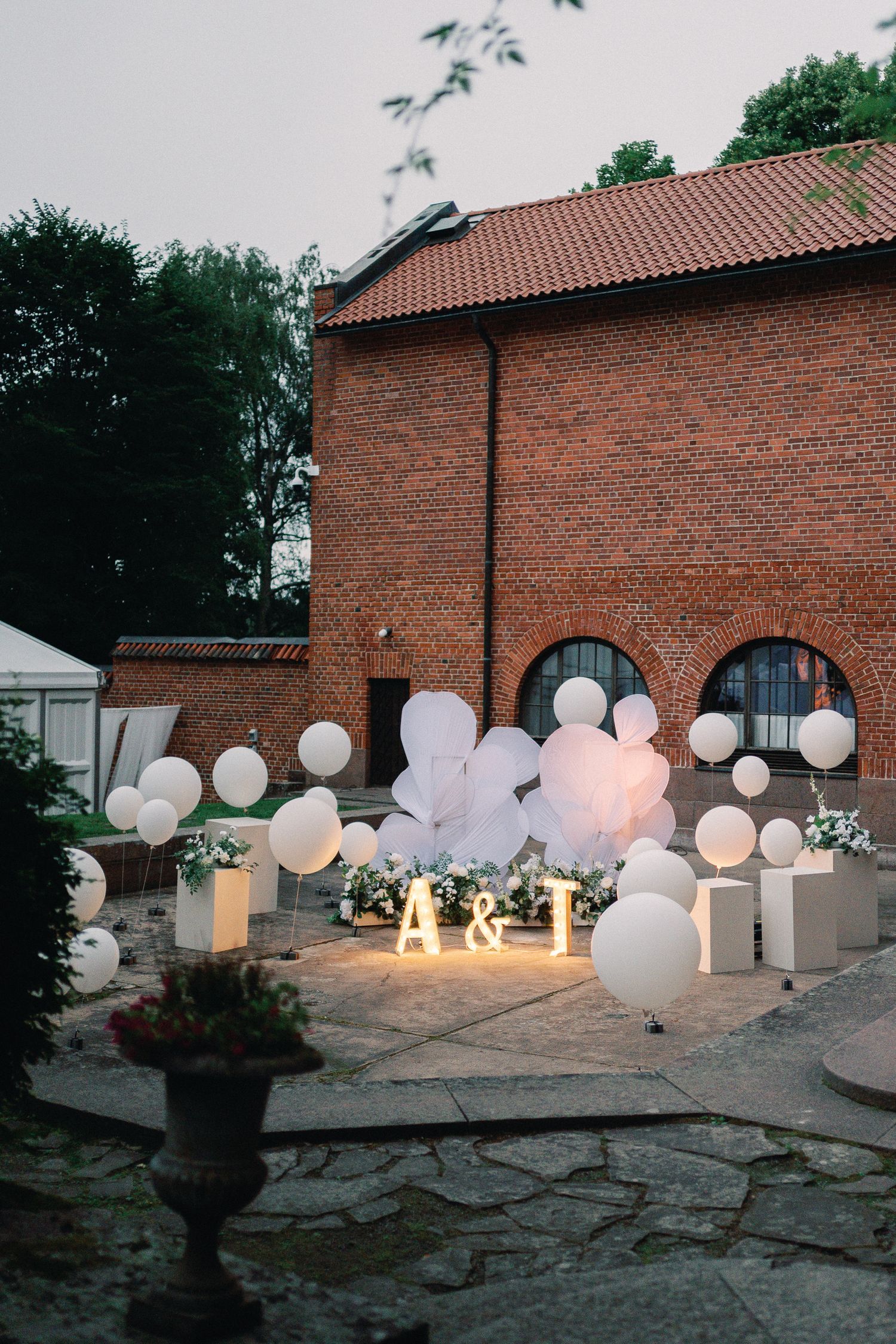 Outdoor wedding decor with white balloons and illuminated A&T letters against a rustic brick building at dusk.