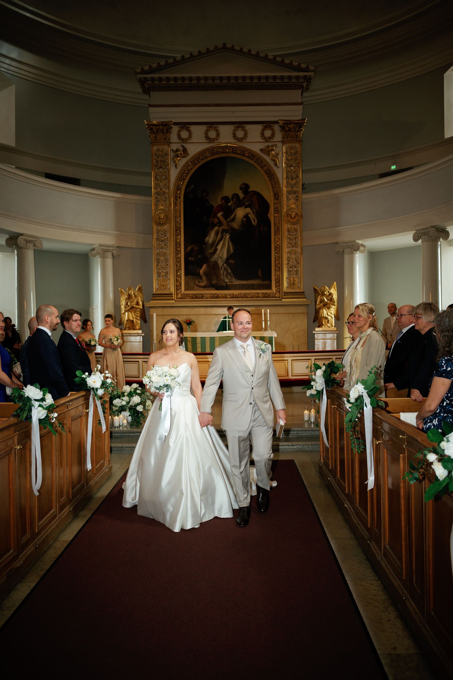 Bride and groom walk down church aisle after wedding ceremony, surrounded by white floral arrangements and wedding guests.