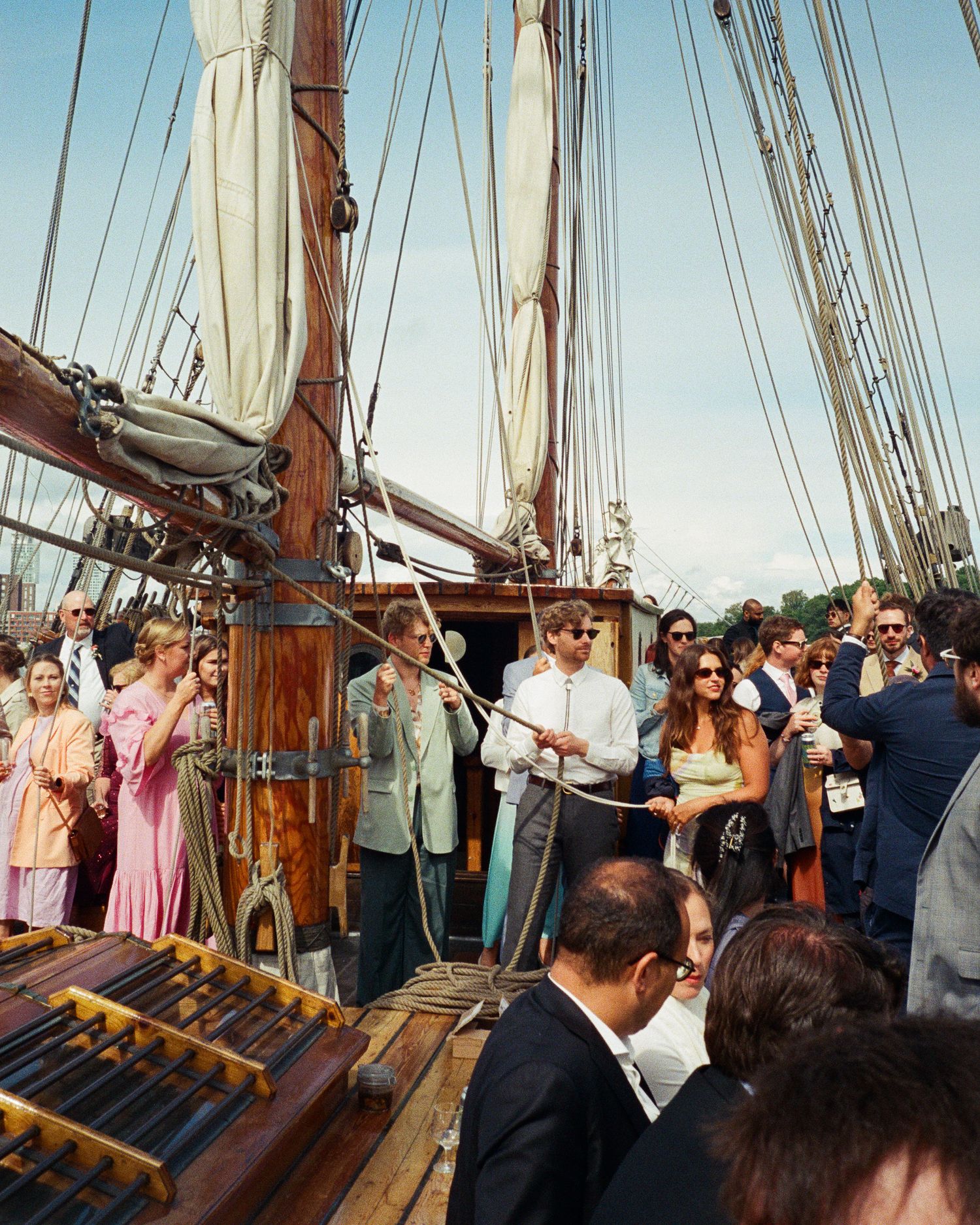 Guests in formal attire gather on the deck of a tall wooden sailing ship during a sunny outdoor event.